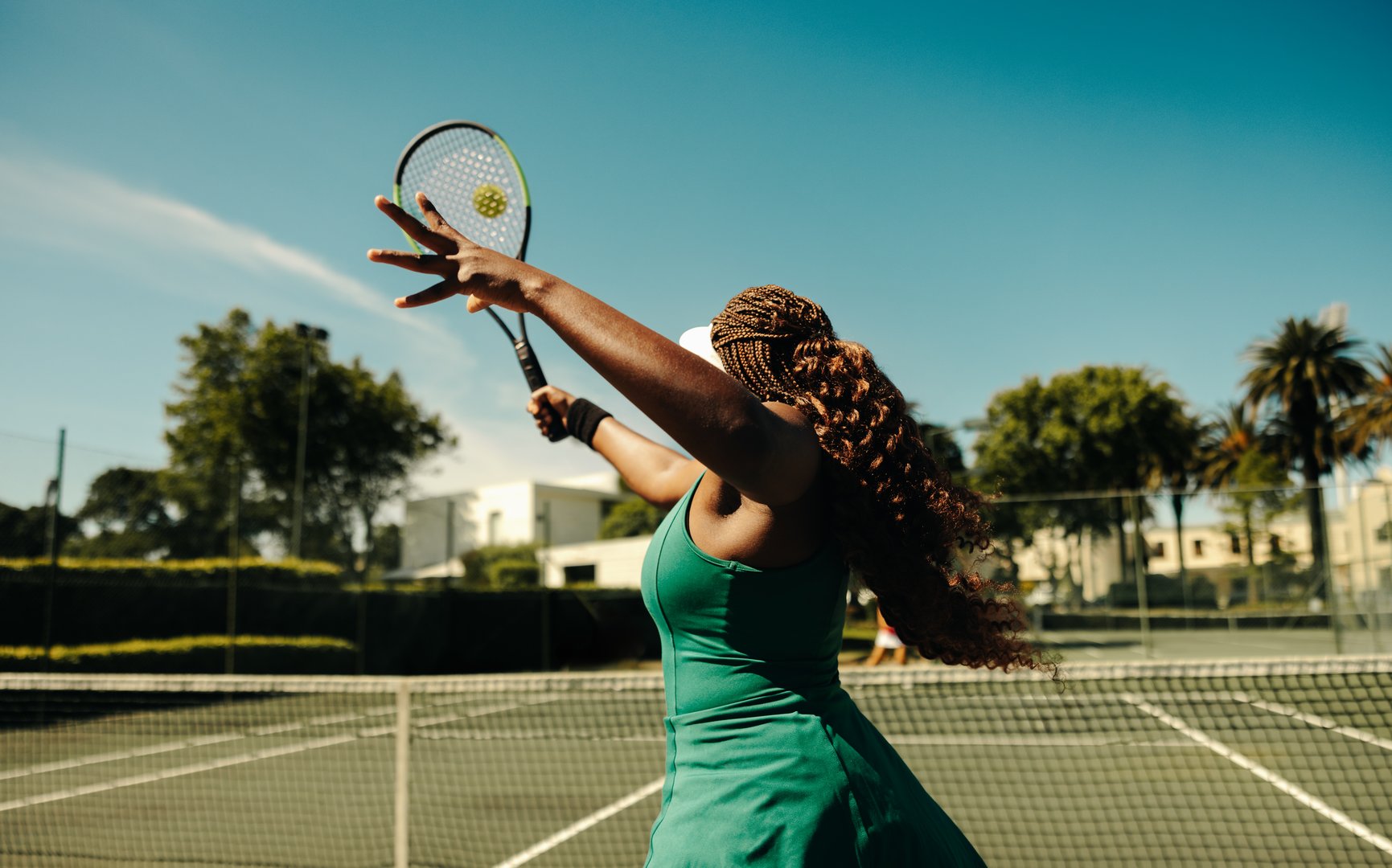 Female tennis player seen from behind, hitting a volley on a hardcourt during a sunny day. Sporty woman playing tennis outdoors.