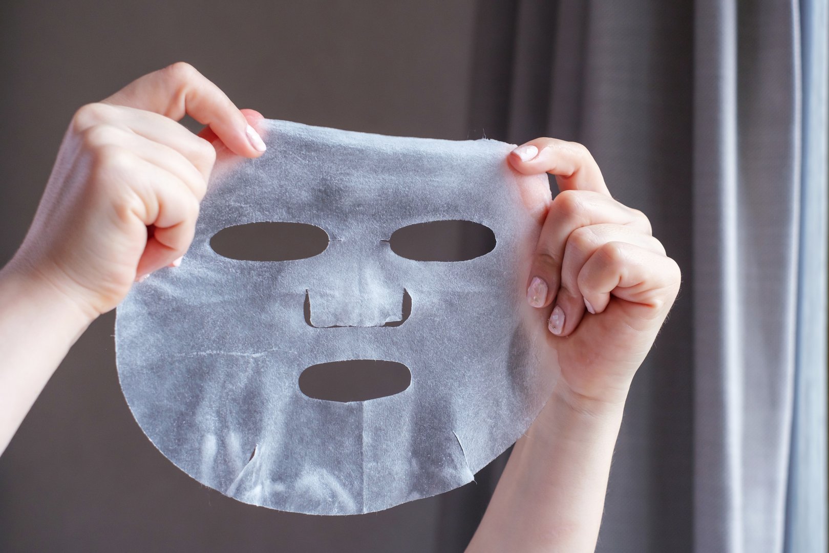 Female hands with white manicure hold fabric mask on a gray background.