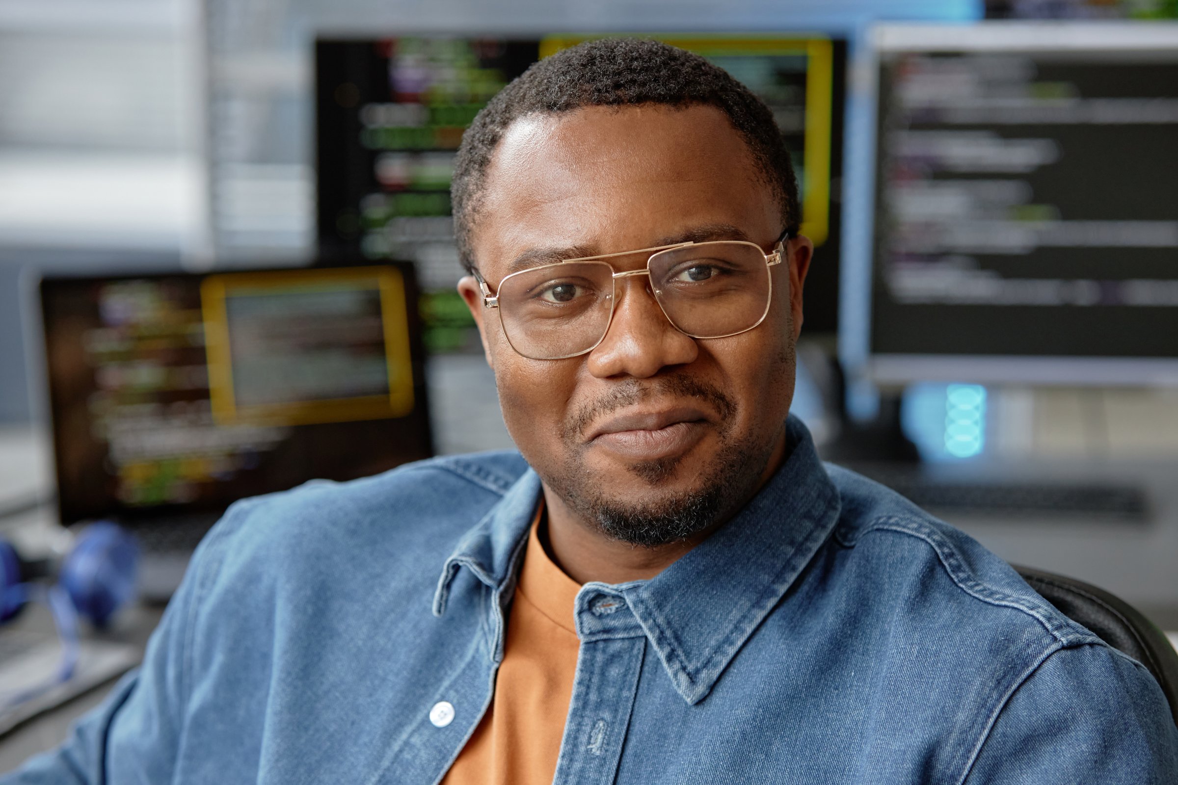 Close up portrait of young male African American VR software engineer in glasses happily smiling looking at camera while sitting in computer programming office, copy space