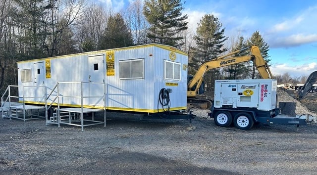 A white portable office with yellow trim on a construction site, next to a generator trailer and an excavator.