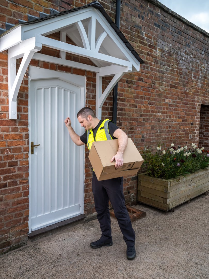A courier in a high-visibility vest knocks on a rural UK brick house door with a package on an overcast day. Ideal for countryside delivery themes.