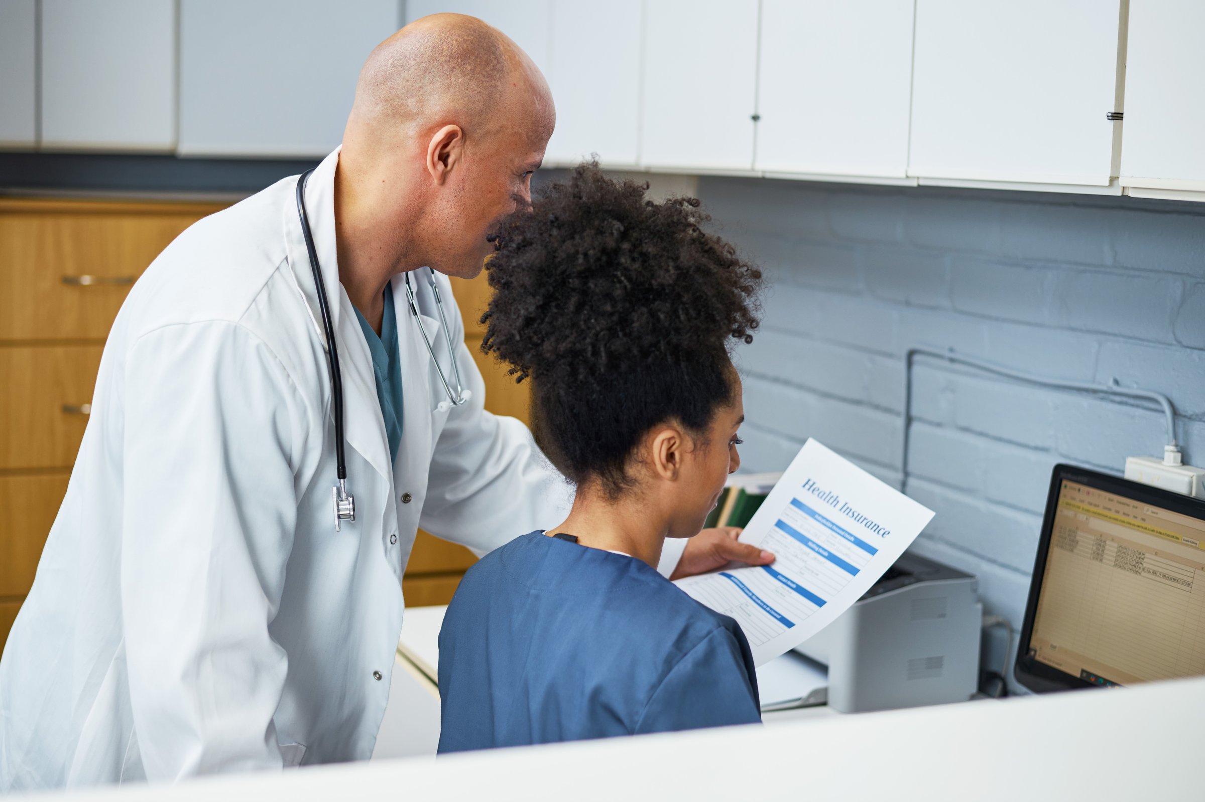 A medical professional reviews health insurance documentation with an assistant in a clinical office. The image evokes concepts of collaboration, healthcare management, and administrative organization in a healthcare setting.