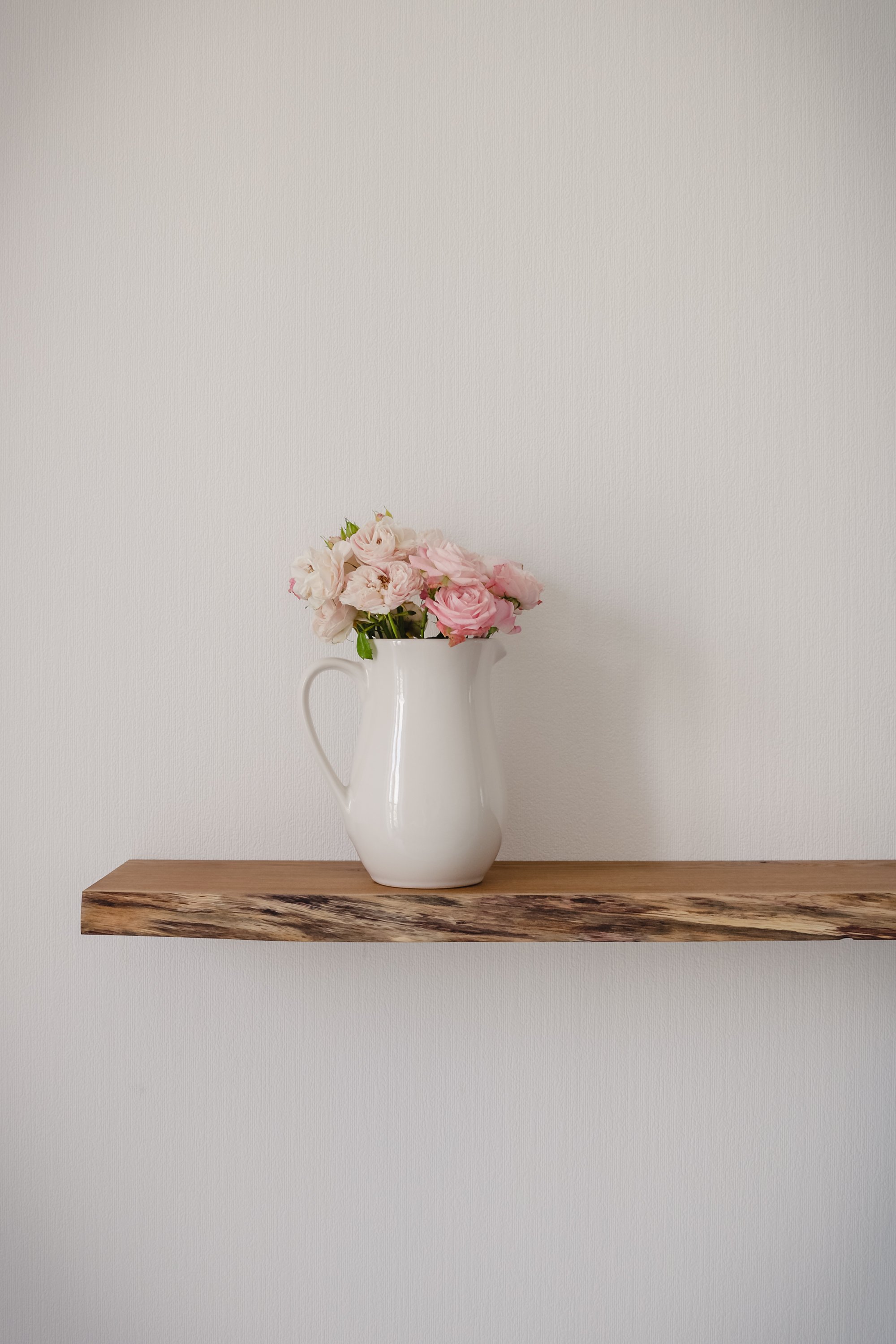 Open floating wooden shelf with a big ceramic jug of pink pastel flowers on a white wallpaper wall in a modern design living room