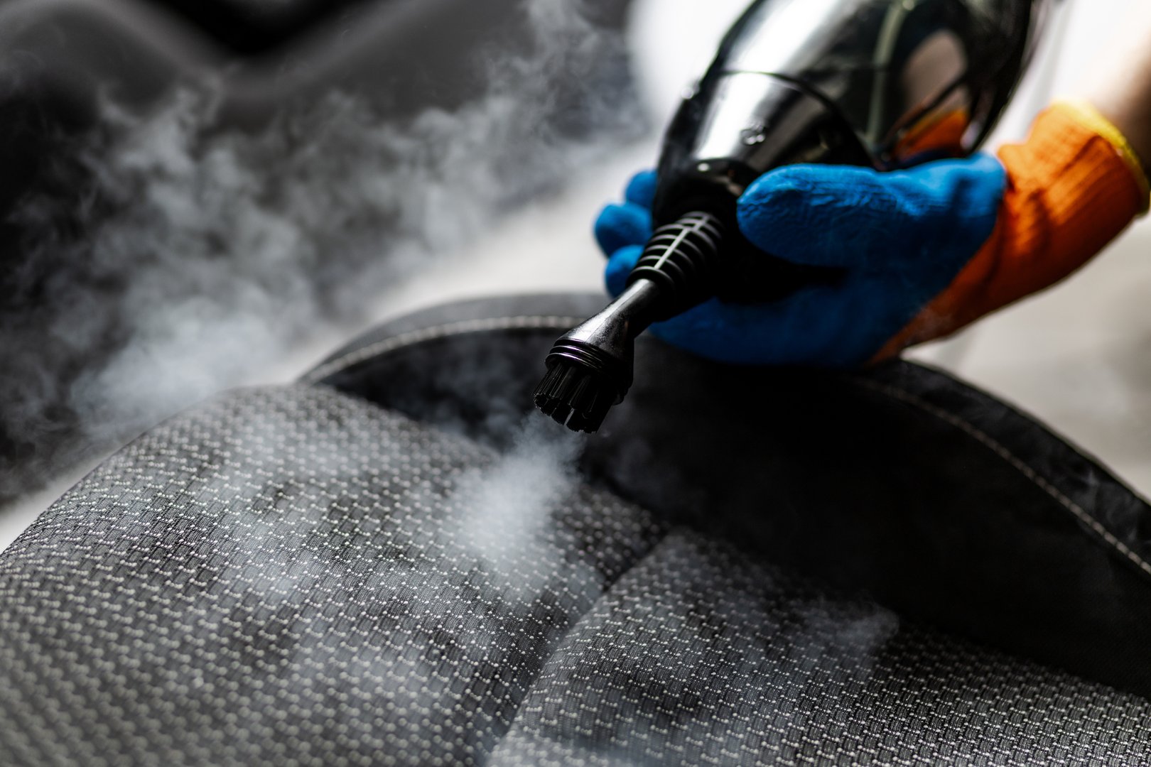A person uses a handheld steam cleaner to deep clean a car seat, releasing steam and targeting dirt and stains efficiently in a well-lit garage.