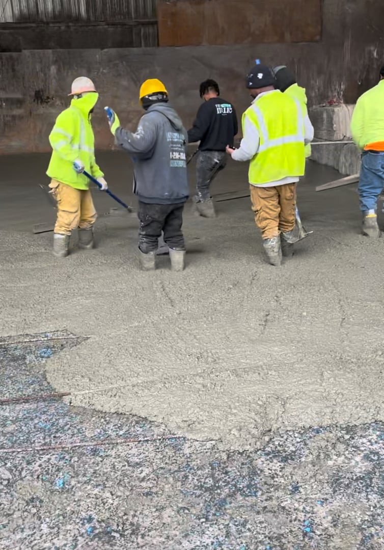 Construction workers smoothing freshly poured concrete on a site, wearing safety gear including boots, helmets, and hi-vis vests.