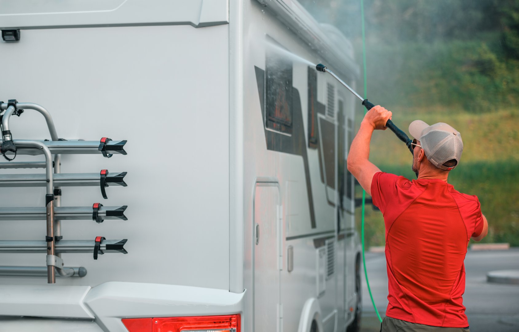 A man in a red shirt washes the side of a white motorhome with a pressure washer. The motorhome has a bike rack on the back and is parked in an outdoor setting.