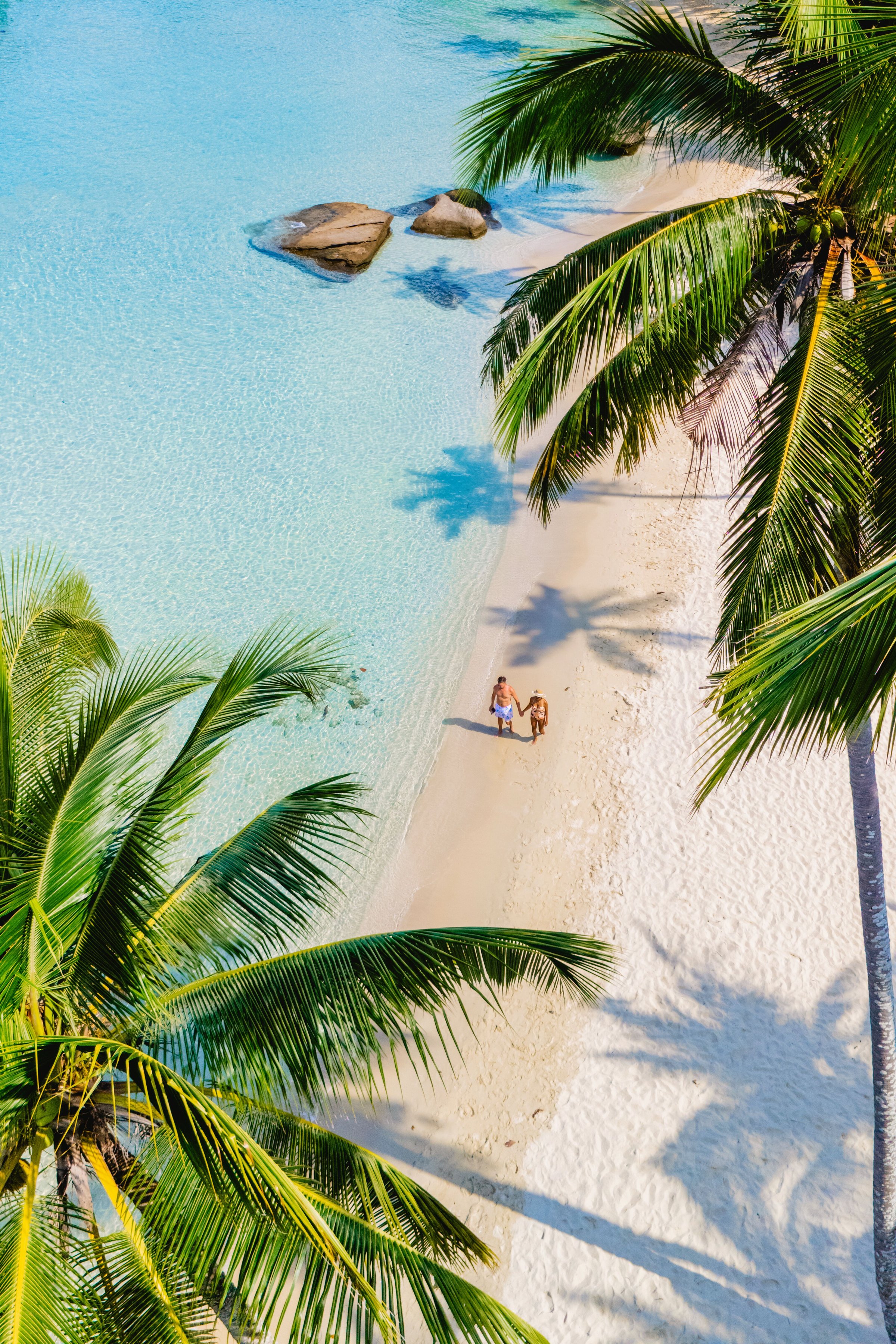 Bright sun casts warm rays as a couple strolling hand-in-hand along a tranquil beach at Koh Kood Thailand. Lush palm trees frame the picturesque scene, highlighting soft white sand and turquoise waves