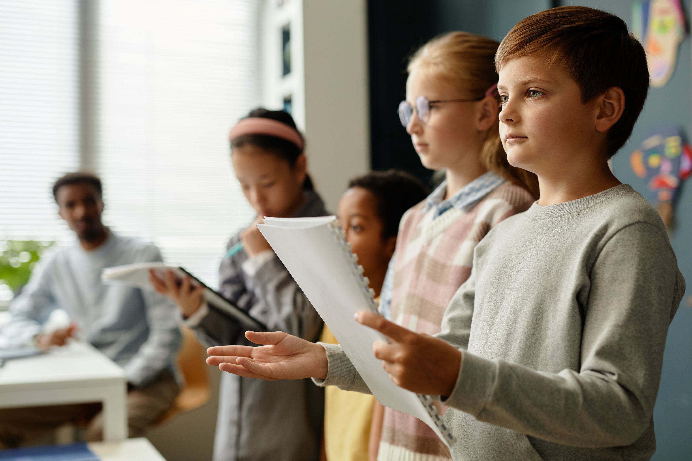 Side view of young students standing in line in classroom and serious looking boy holding exercise book presenting homework during lesson at school, copy space