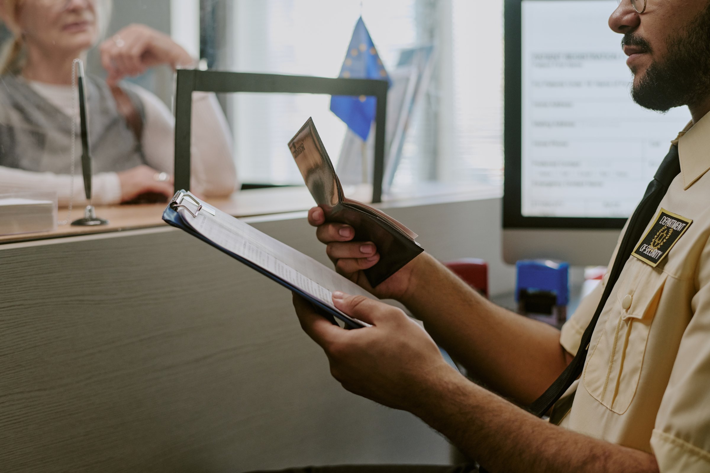 Person at immigration desk reviewing documents and passport. Handing over paperwork with background of computer screen and another individual