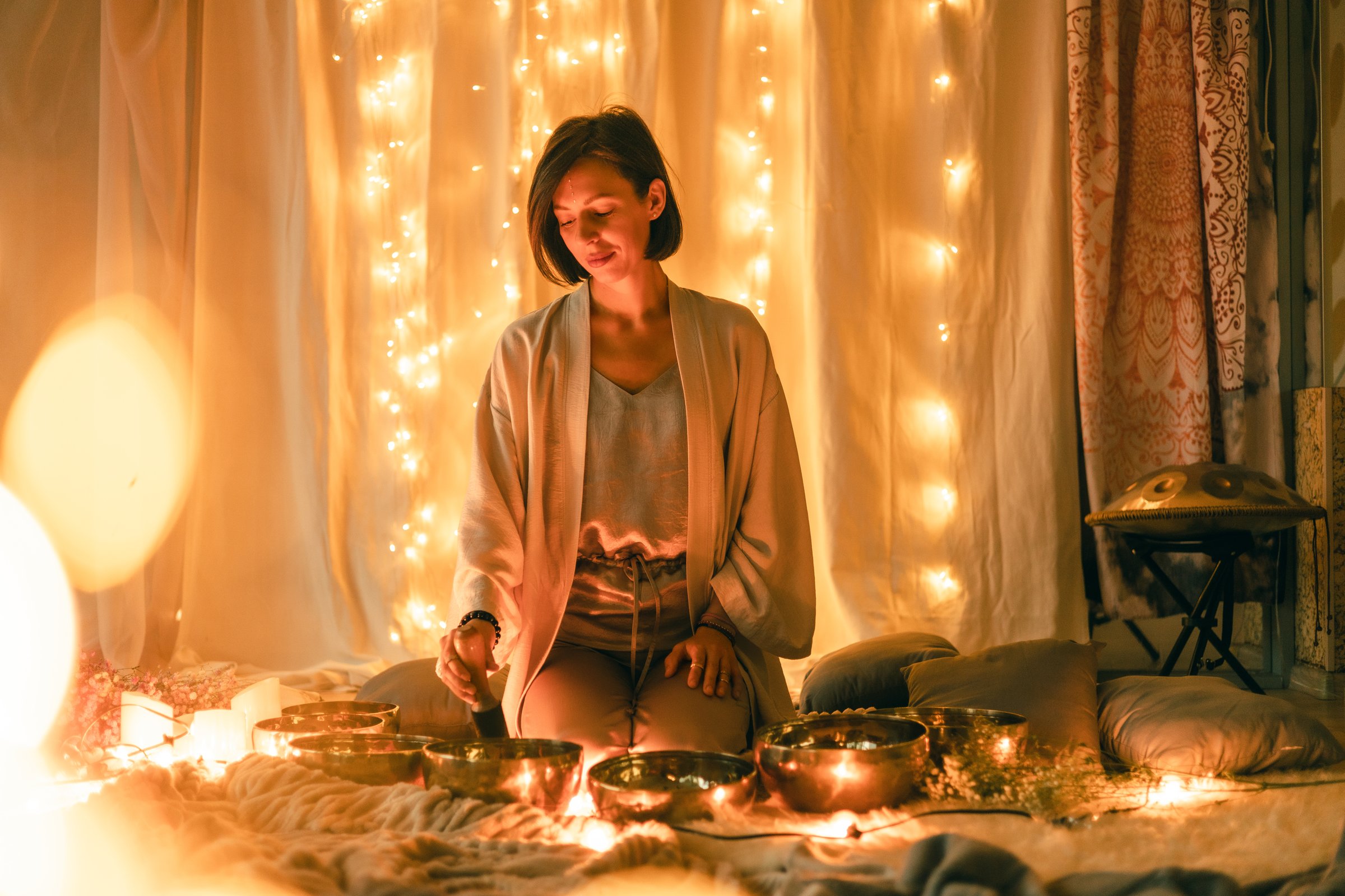 Woman sitting among singing bowls in a softly lit meditation space, surrounded by warm lights and calming decor, creating a peaceful atmosphere for relaxation, mindfulness, and sound healing practice.