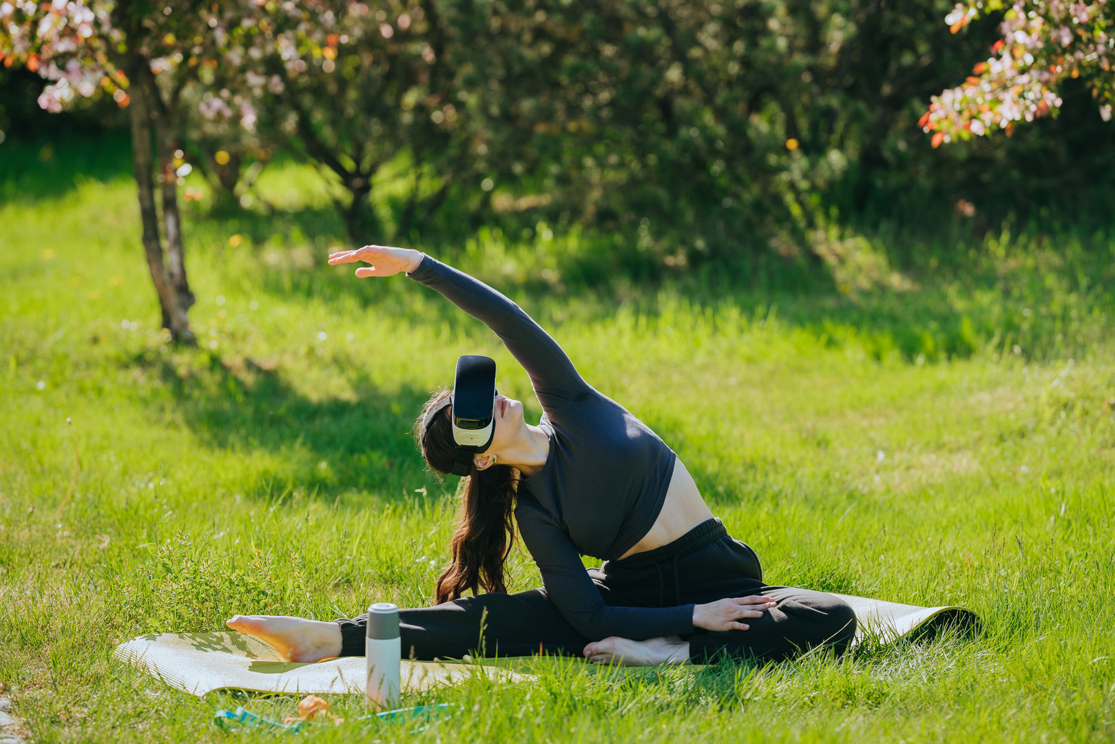 Woman enjoys an innovative outdoor yoga session, wearing a VR headset on a lush lawn, blending technology with relaxation under flowering trees.
