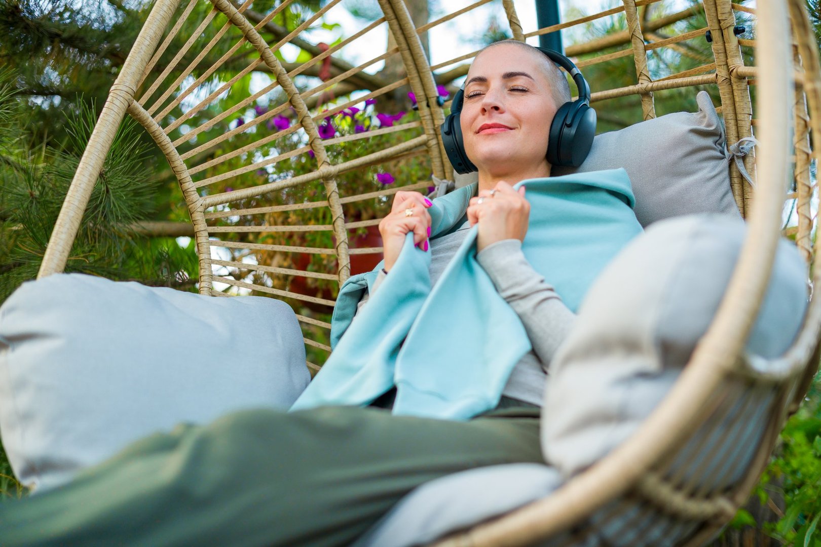 Young female cancer patient relaxing in hanging chair on the garden patio, listening to music. Mental wellbeing during illness.