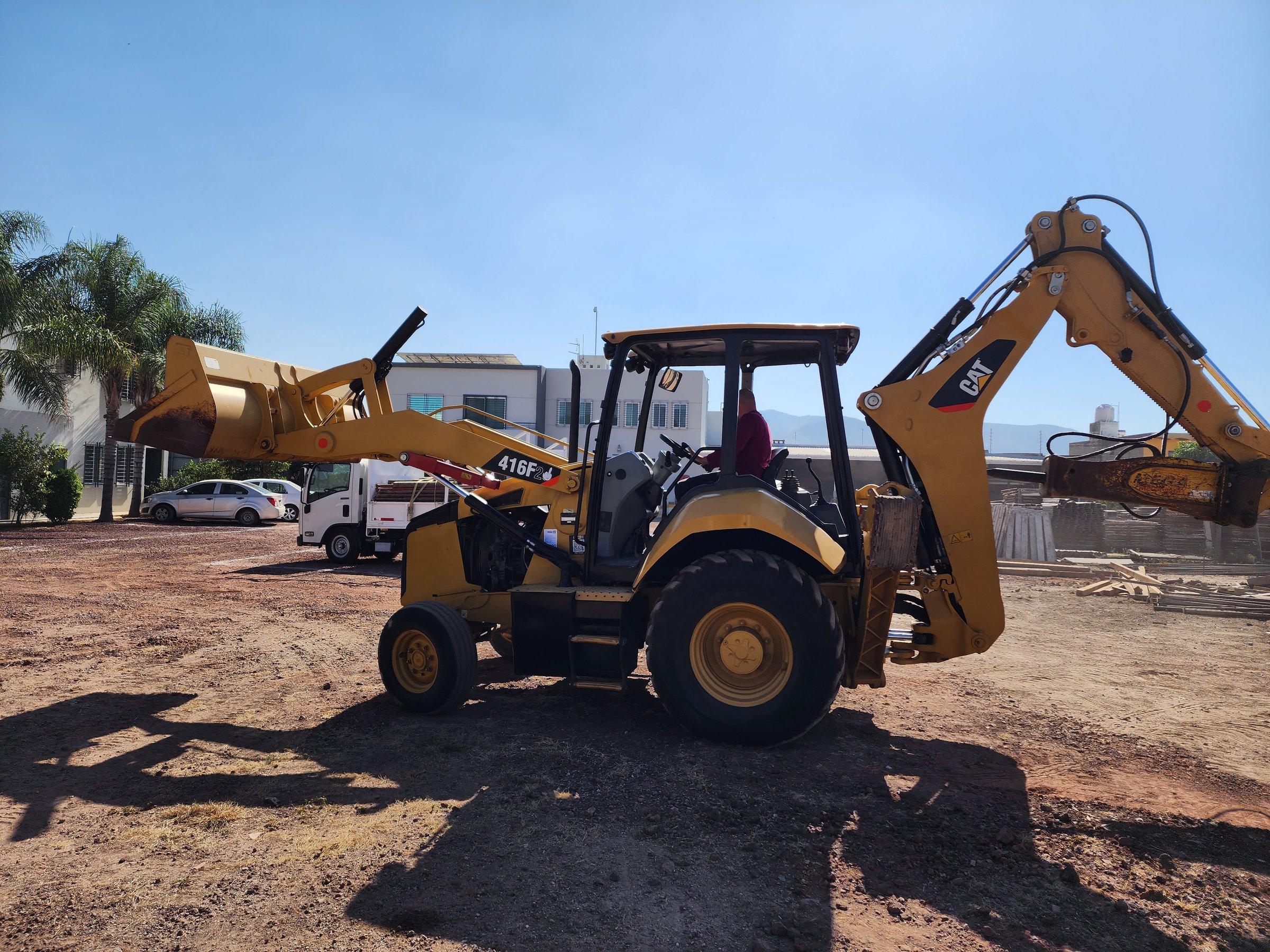 A yellow backhoe loader parked on a construction site with buildings and trees in the background under a clear blue sky.