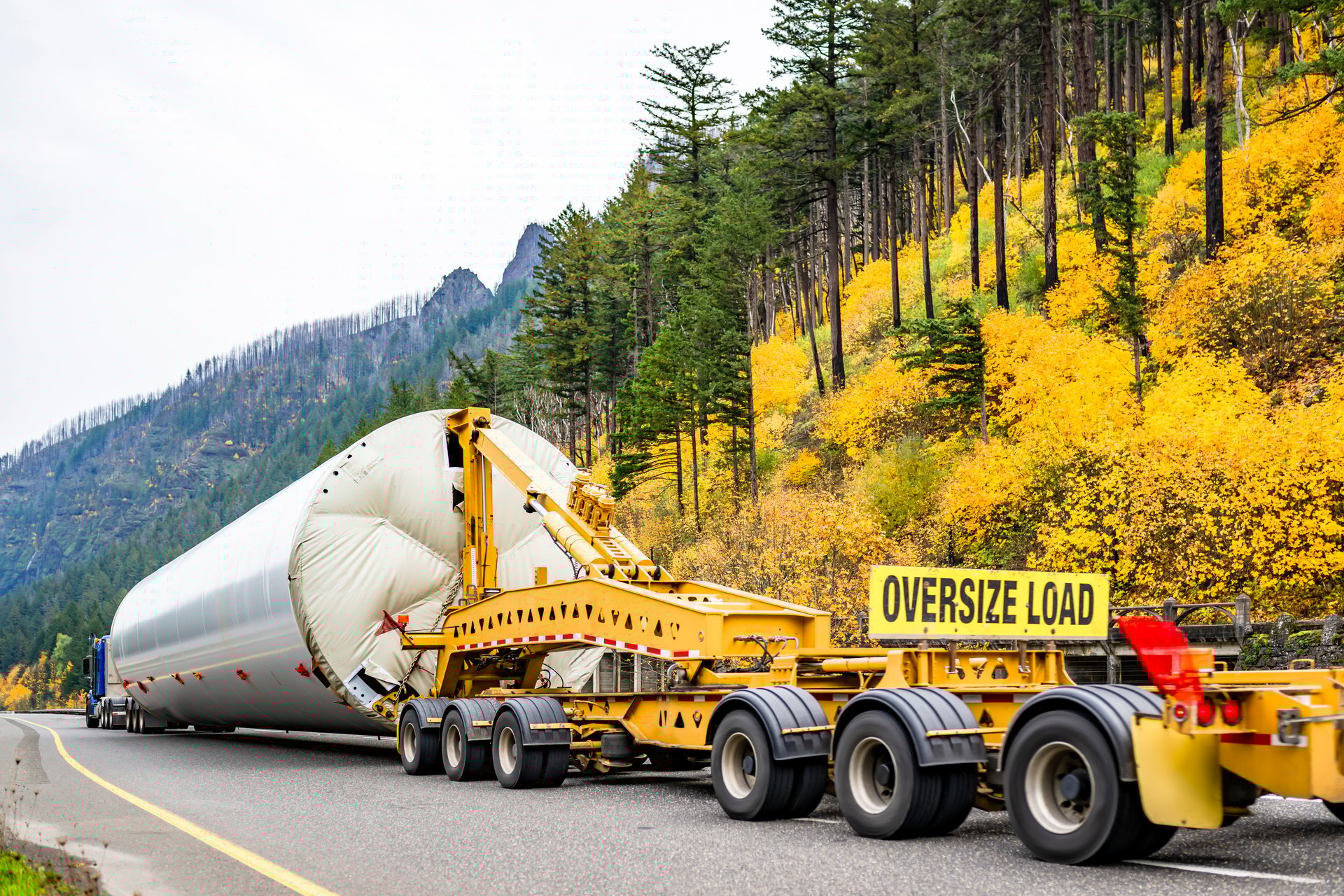 Big rig semi truck transporting oversized part of the wind generator support pole using an additional heavy duty trolley with six axles and oversize load sign driving on the autumn highway road