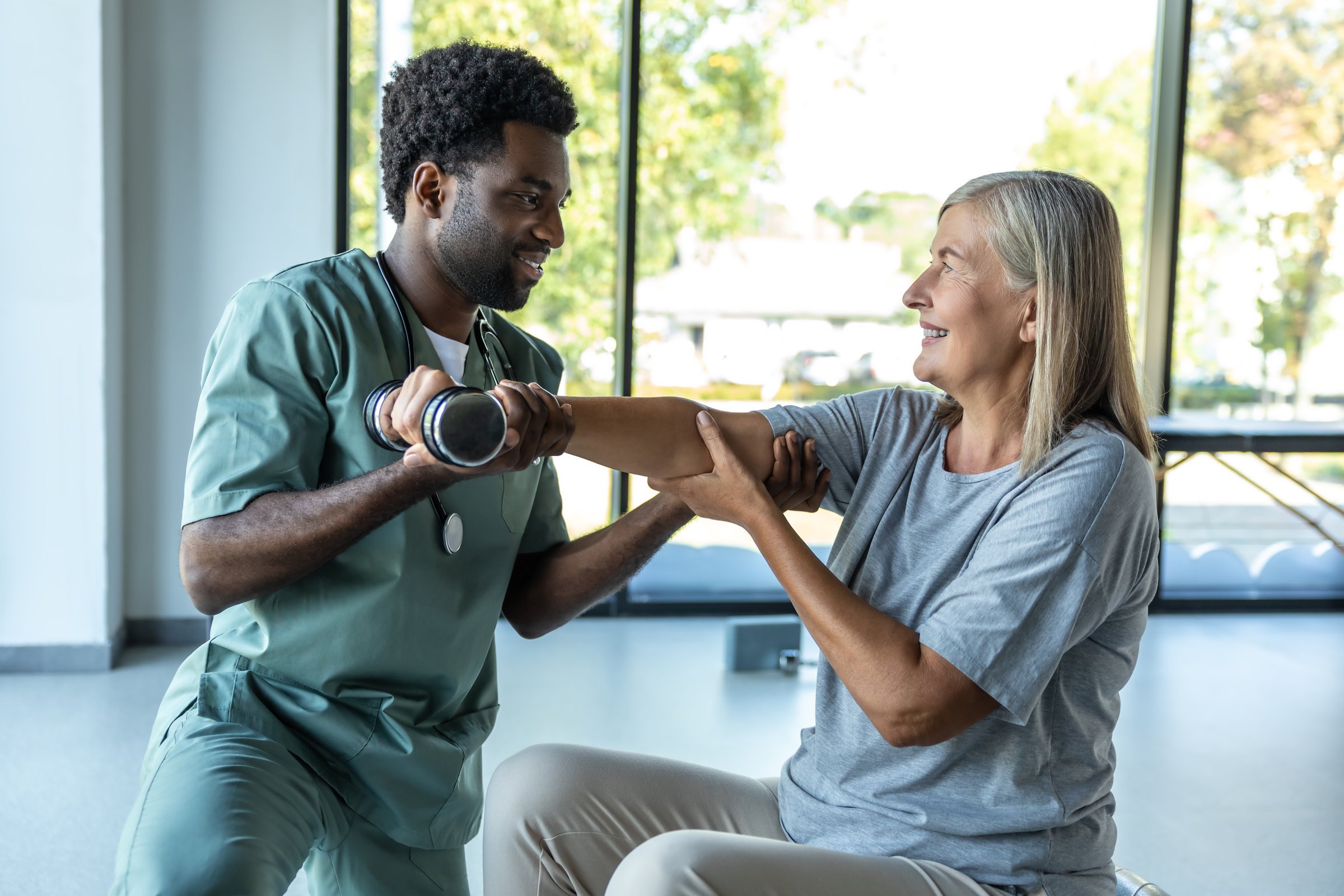 Man doctor providing physiotherapy and rehabilitation with dumbbells to adult woman patient for recovery in clinic