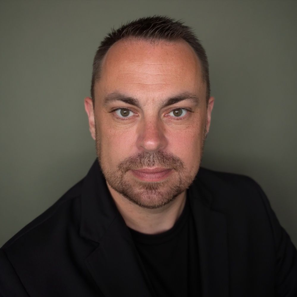 Man with short dark hair and a beard, wearing a black blazer and shirt, looking directly at the camera against a grey background.