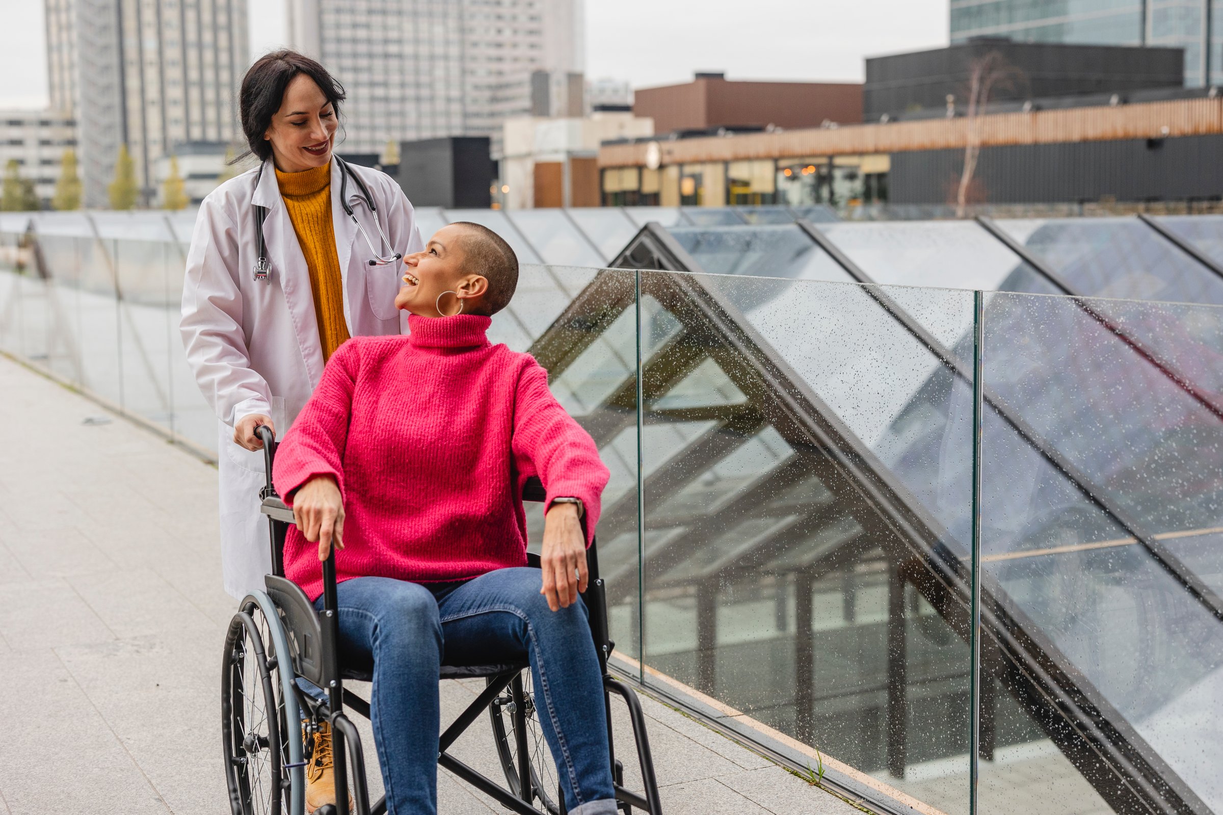 A moment of genuine connection as a smiling doctor communicates with her patient in a wheelchair against an urban backdrop.