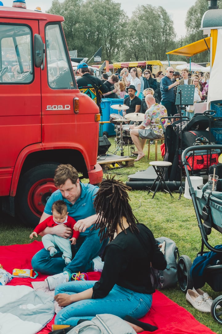Westerpark, Amsterdam, Netherlands: May 20 2023: Young family with little boy enjoys a picnic at Rollende Keukens food festival sitting on a blanket on a grass in the midst of food stalls and cars