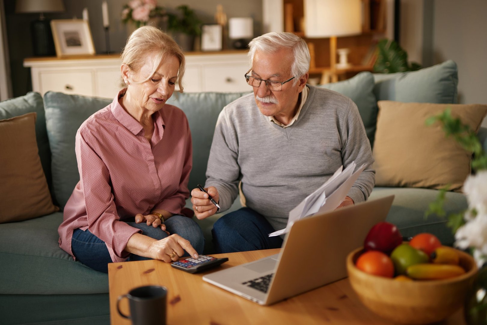 Senior couple using a calculator and laptop, managing their home finances and paperwork together on the sofa