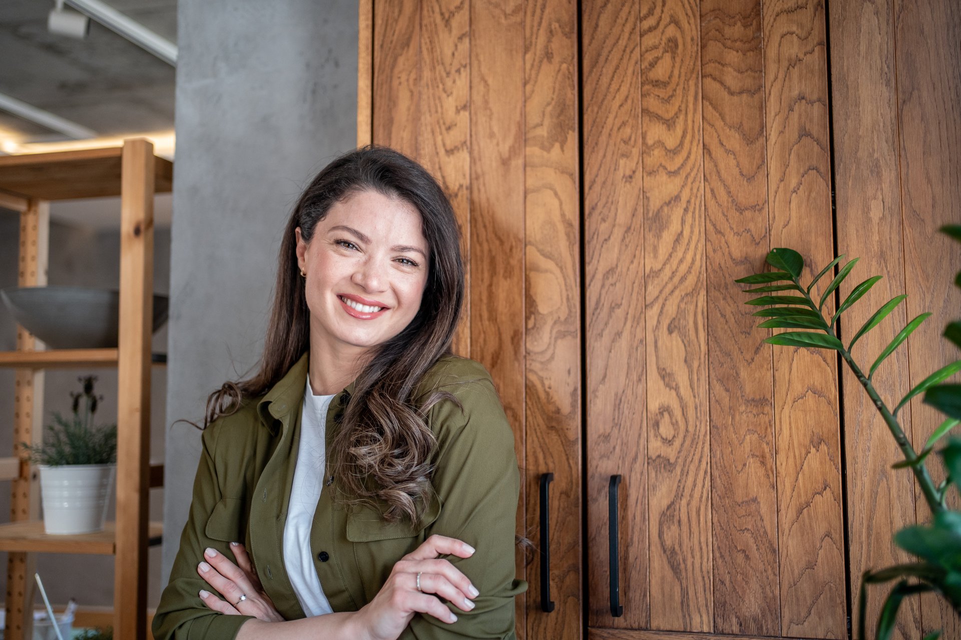 Young businesswoman with crossed arms is smiling and leaning against a wooden cabinet in a modern office, exuding confidence and professionalism