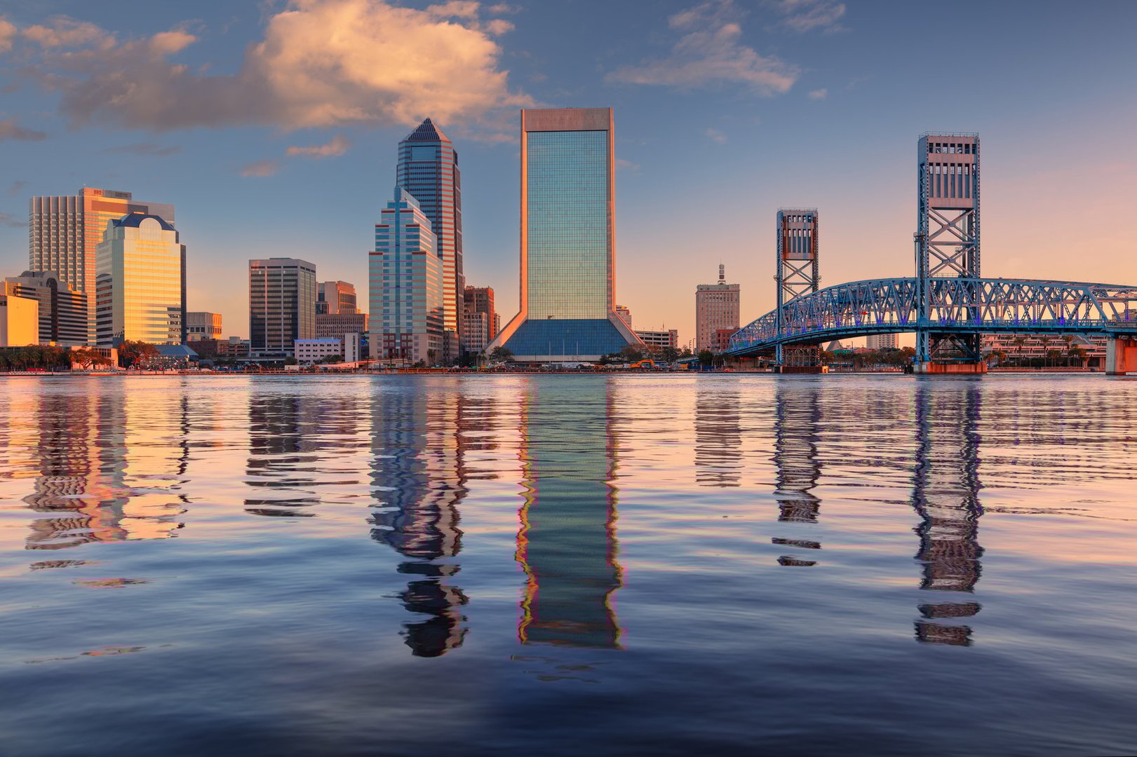 Skyline of Jacksonville, Florida at sunrise, featuring modern high-rise buildings reflecting in the calm water of the St. Johns River. The scene includes a prominent blue truss bridge on the right, adding architectural interest. The sky is painted with hues of orange and pink, with scattered clouds accentuating the warm glow of the early morning sun. These elements create a serene and picturesque urban landscape.