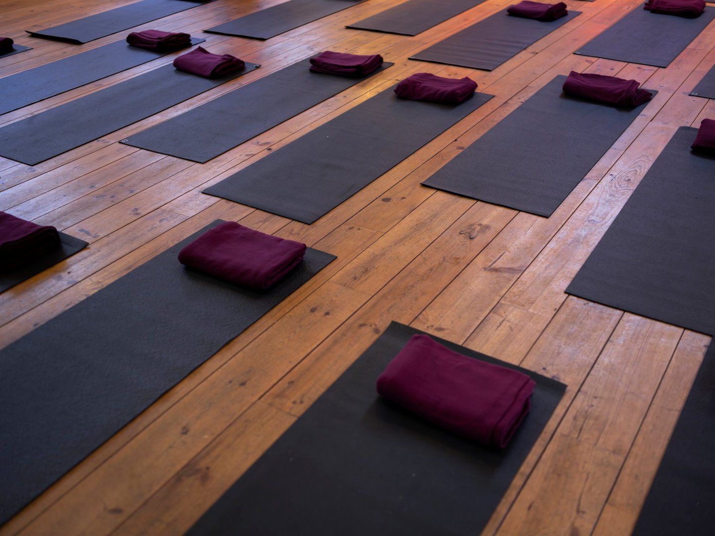 A serene yoga studio is shown with multiple mats neatly laid out on a wooden floor, each with a rolled purple towel. Warm orange lighting illuminates the space, highlighting the architectural details of the historic building, including large windows and pillars. A grand piano and floral arrangement add to the peaceful atmosphere.