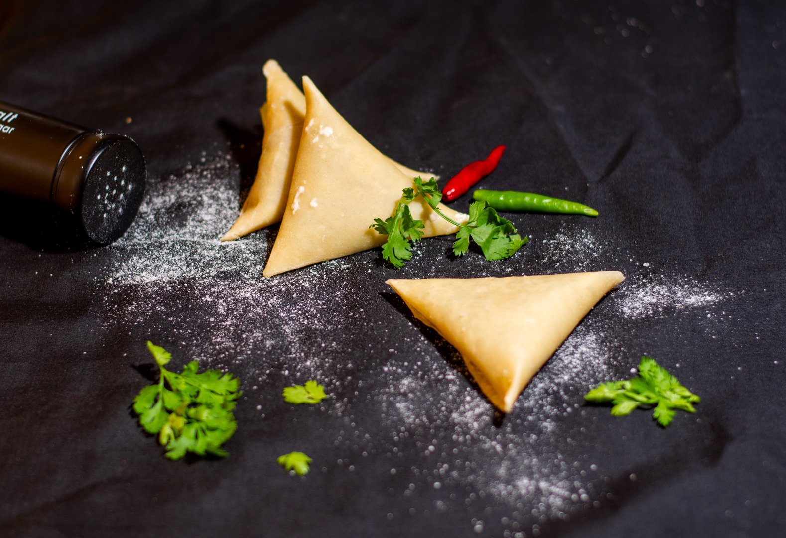 Three samosas on a black surface, sprinkled with flour, garnished with cilantro and two chili peppers.