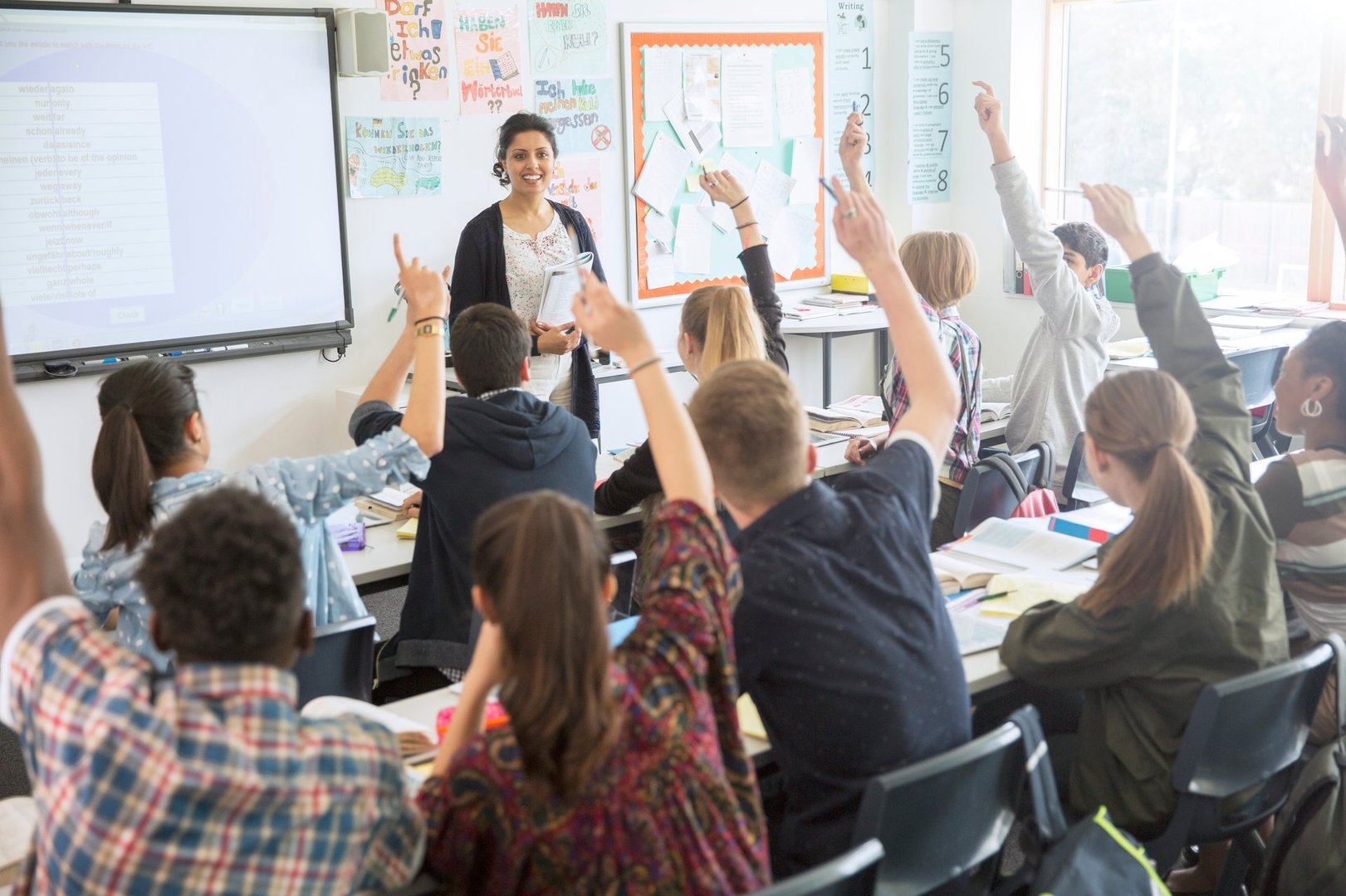 A teacher standing in front of a classroom with students raising their hands during a lesson.