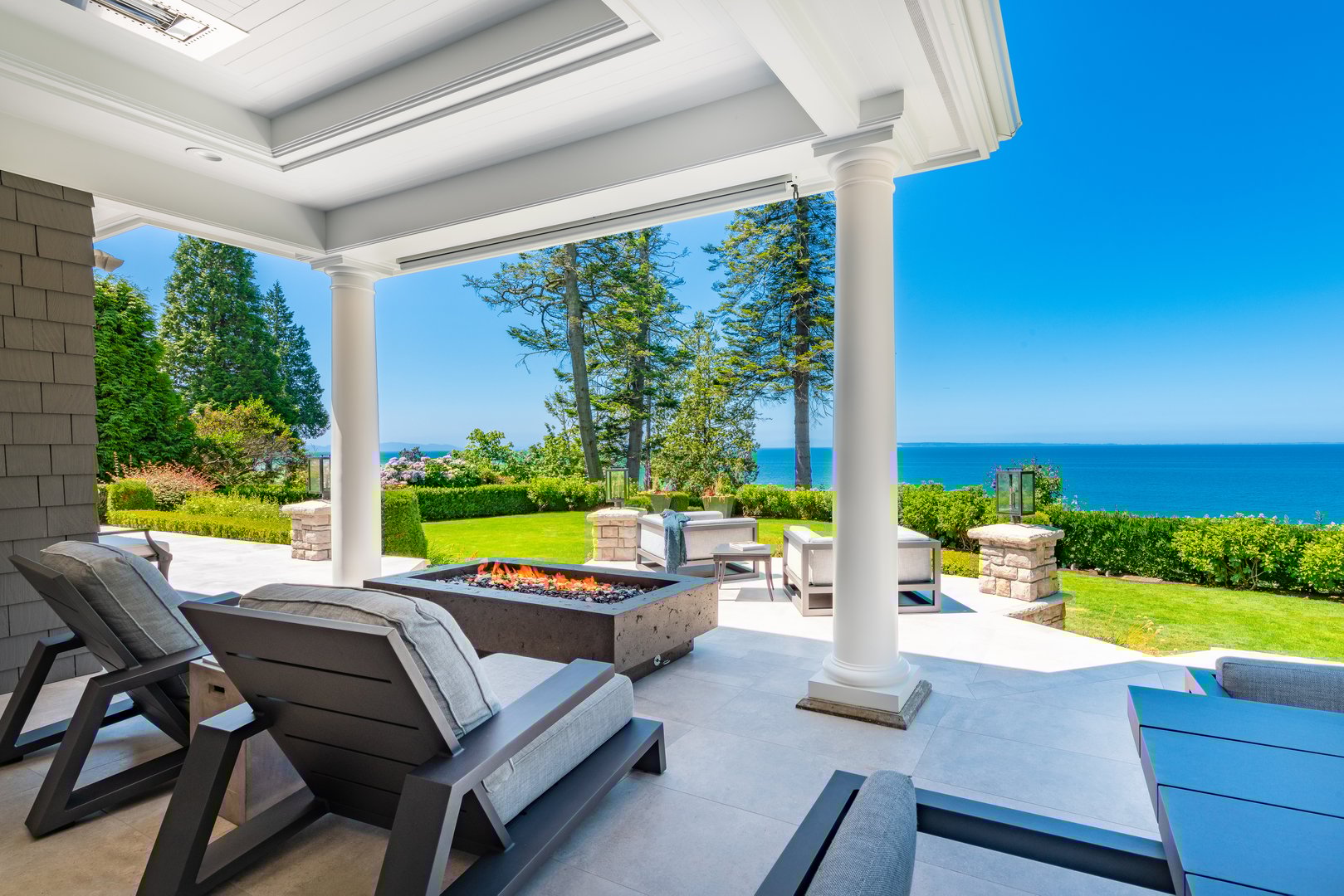 Photographs of the interior and exterior of a beautifully estate home in the Pacific Northwest, this home has been completely remodelled and is a waterfront home with vast views of the pacific ocean and san juan islands in the distance. Interiors show a grand entry foyer and staircase with wrought iron rails. Some interior views of the dining room with a banquette window seat looking over the sea and the patio contains outdoor furniture a fire pit and hot tub hot tub overlooking the grassy yard on a bright sunny day in July.