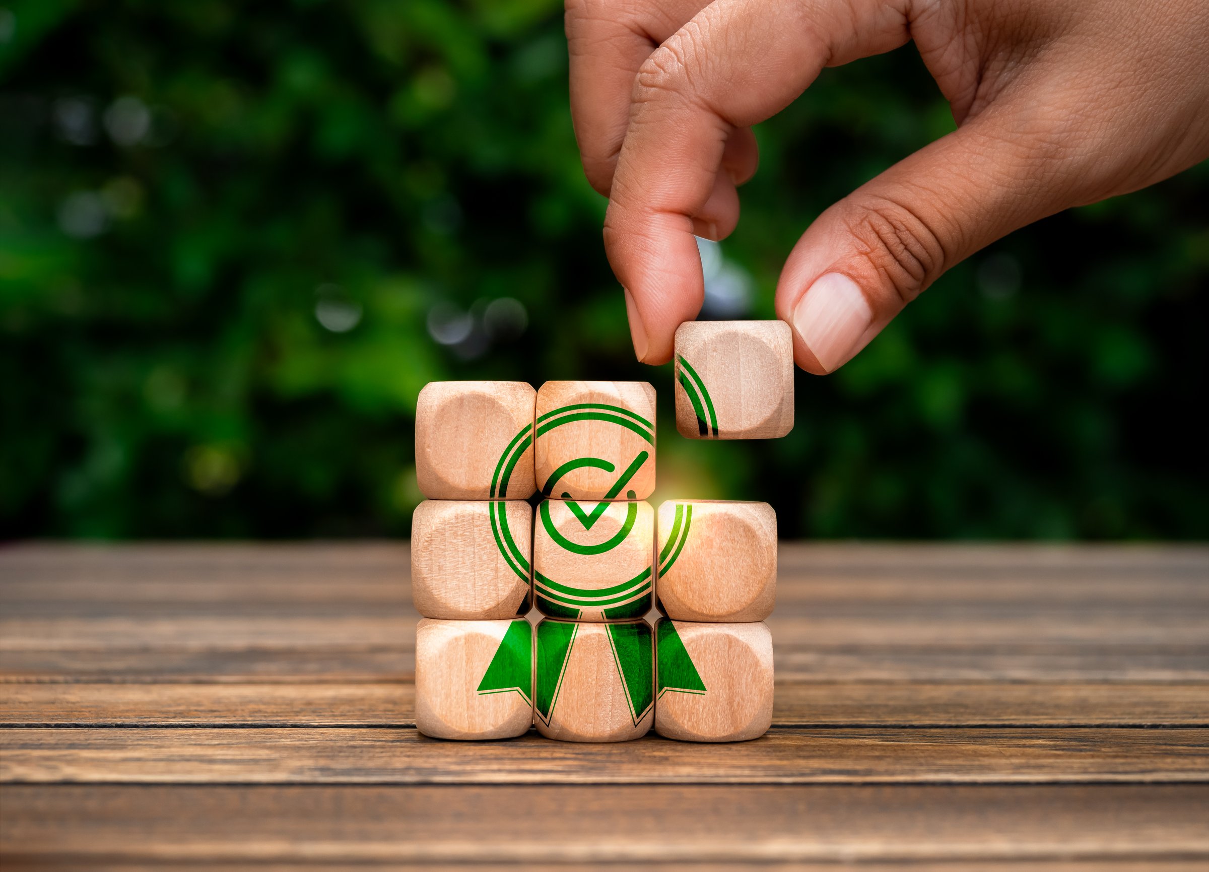 Business quality and service success concept. Hand put last piece of small wooden cube block to completed on puzzle stack with big green warranty icon on wood table on natural green leaves background.
