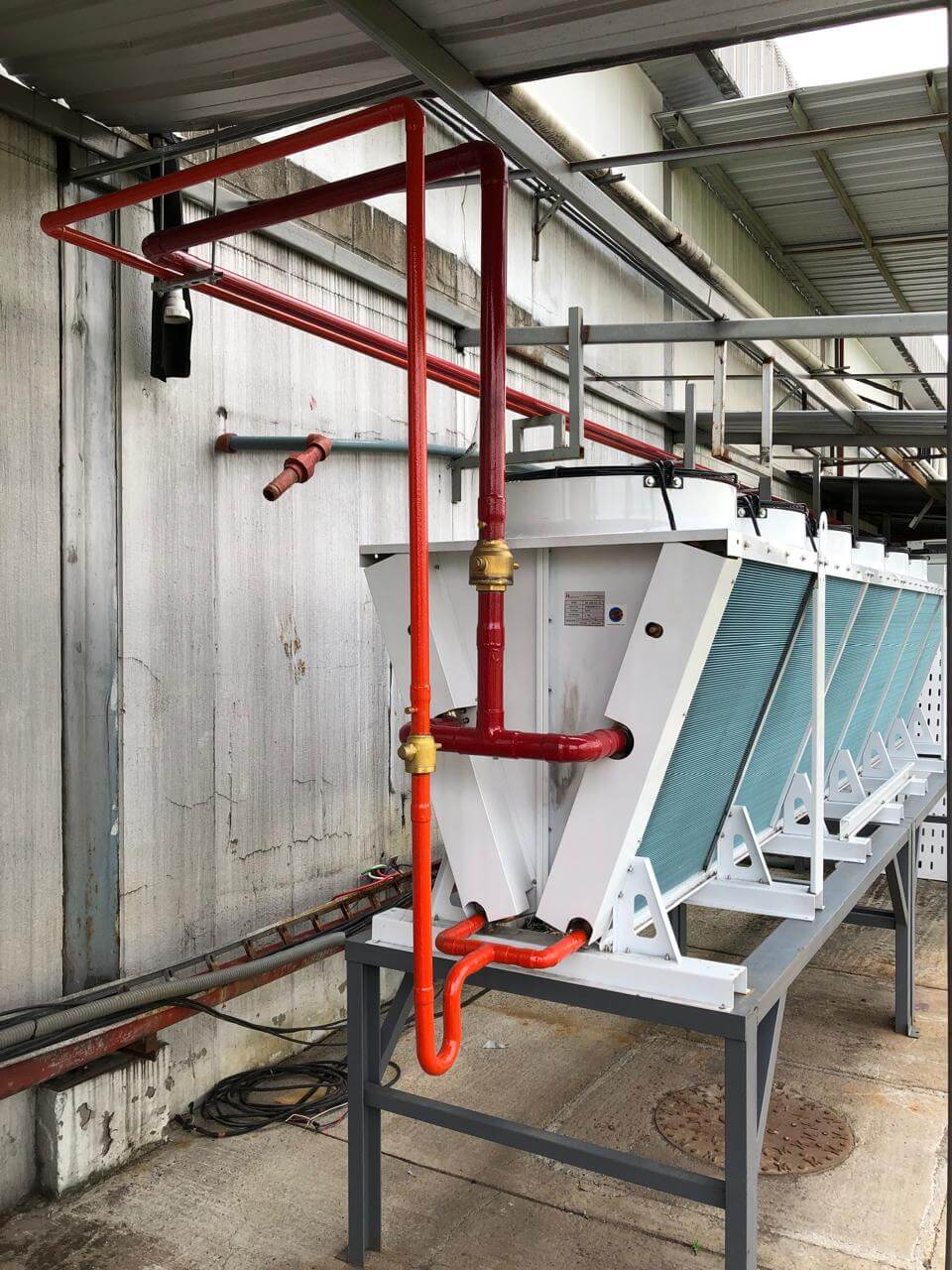 Outdoor industrial cooling unit with red and orange pipes on a metal framework beneath a corrugated roof.