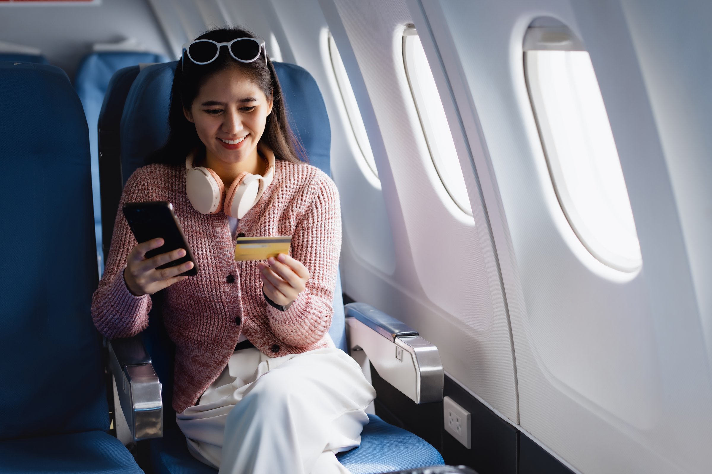 An Asian woman smiles happily on an airplane while using her credit card and smartphone, managing financial transactions, booking travel tickets, or enjoying online shopping during her journey