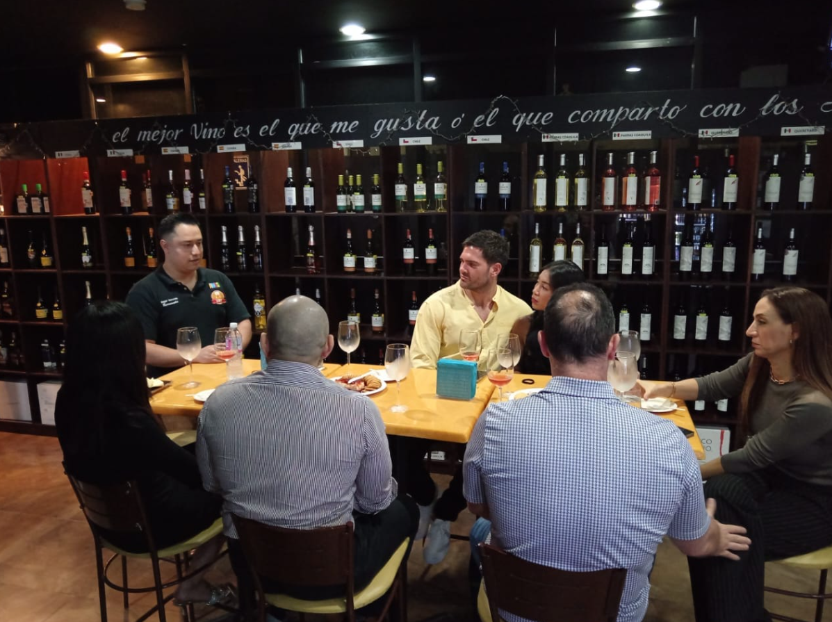 Group of people seated around a table in a wine shop, conversing and enjoying drinks. Wine bottles line the shelves behind them.