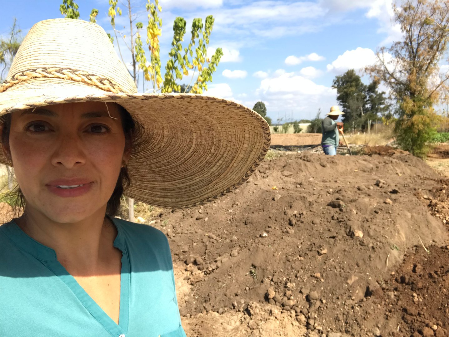 A woman in a wide-brimmed hat takes a selfie on a sunny day in a garden, with another person working in the background.