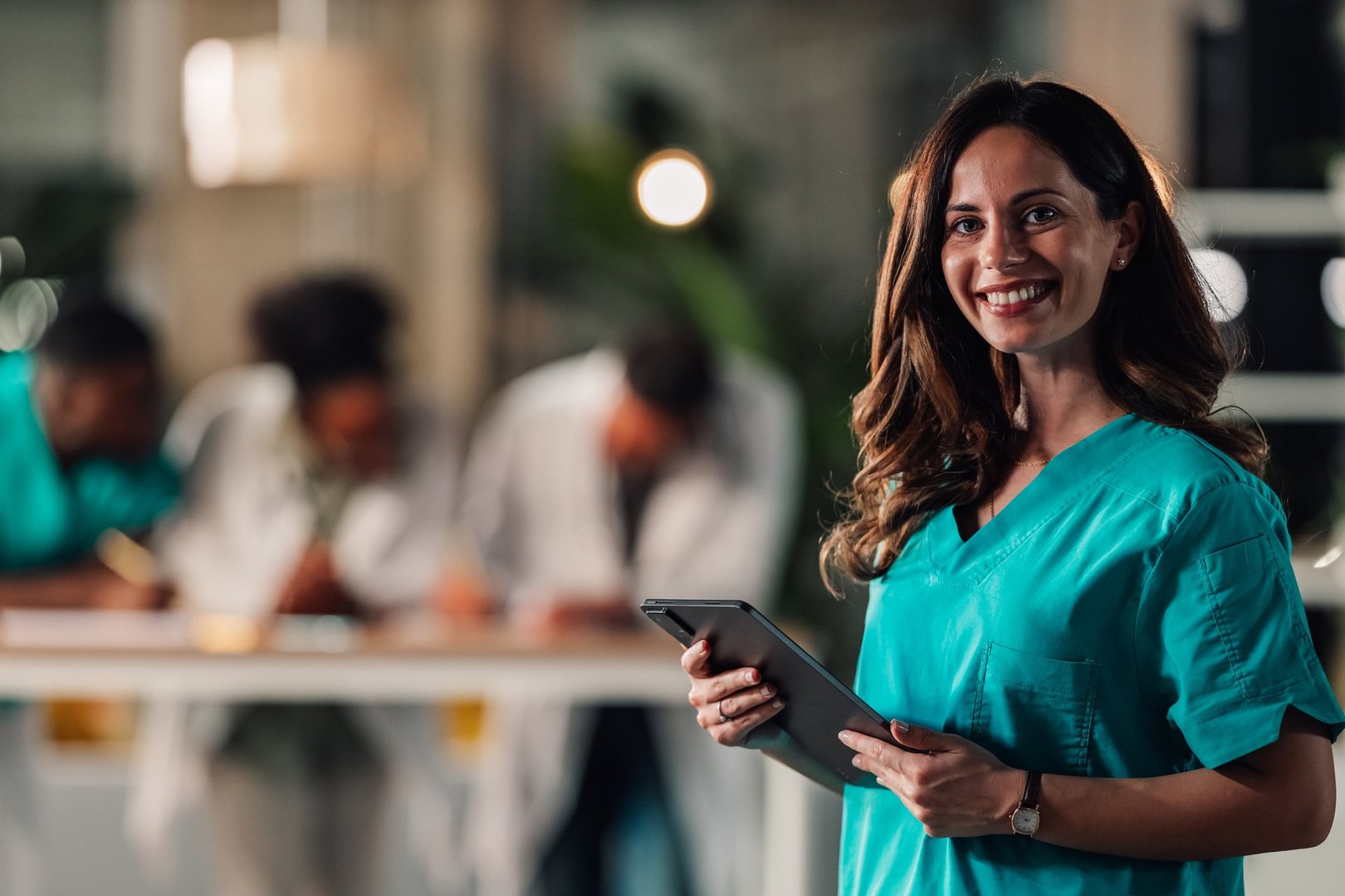 Female doctor or nurse in scrubs smiling, holding a digital tablet with medical team working in background