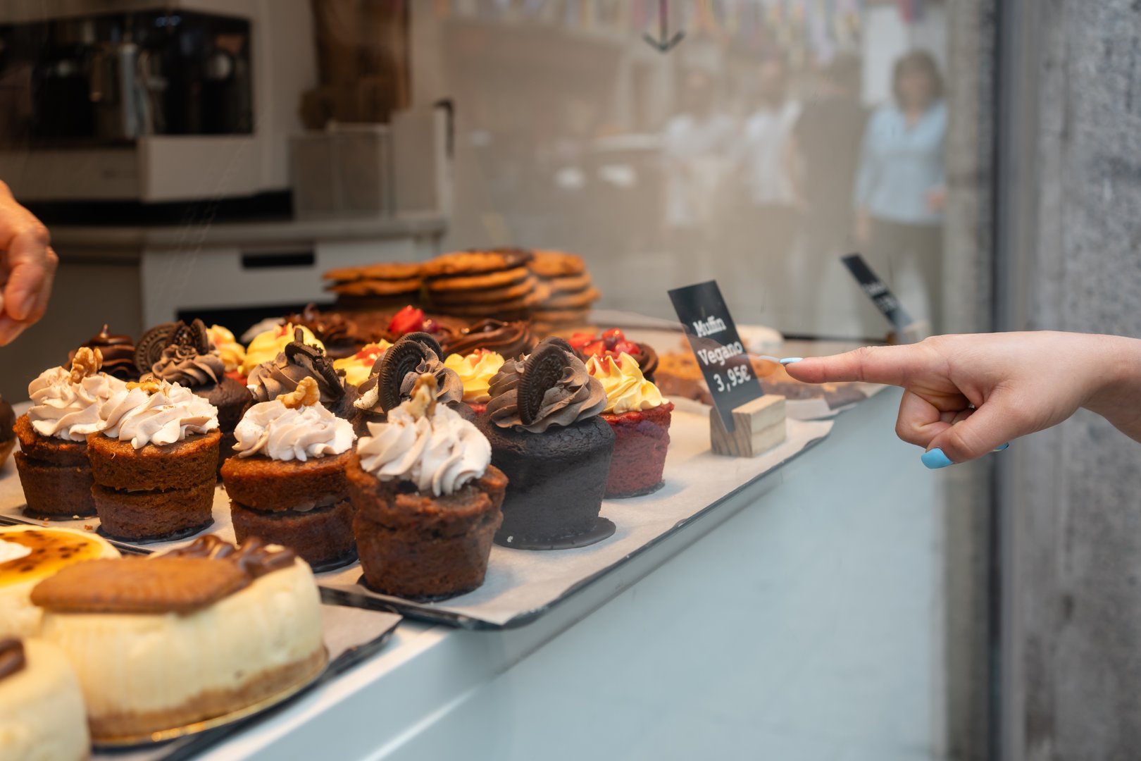 Variety of delicious cupcakes are displayed in a bakery window, with a woman pointing at a vegan muffin