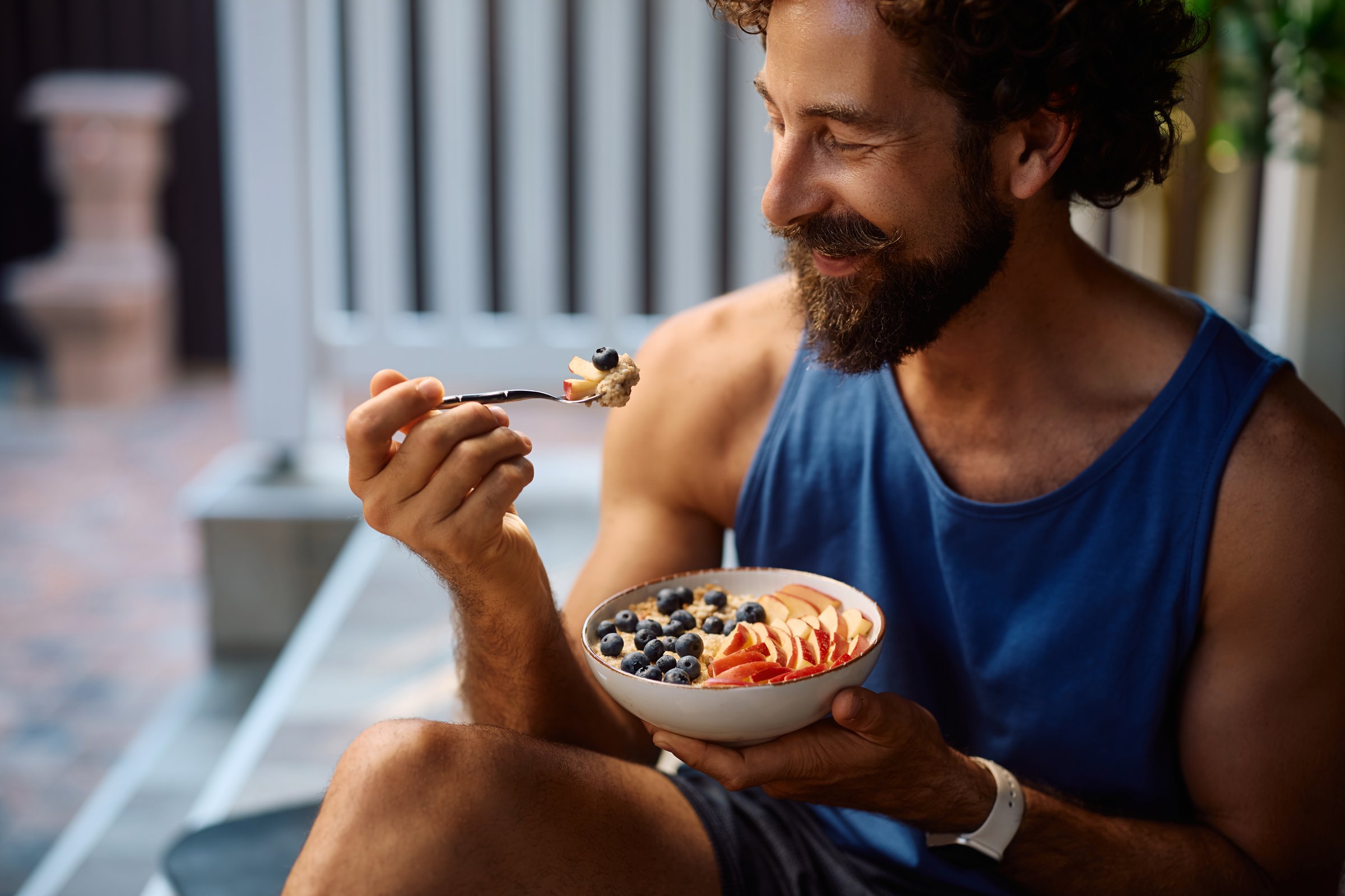Happy sportsman enjoying in fruit oatmeal after working out on a patio at home.