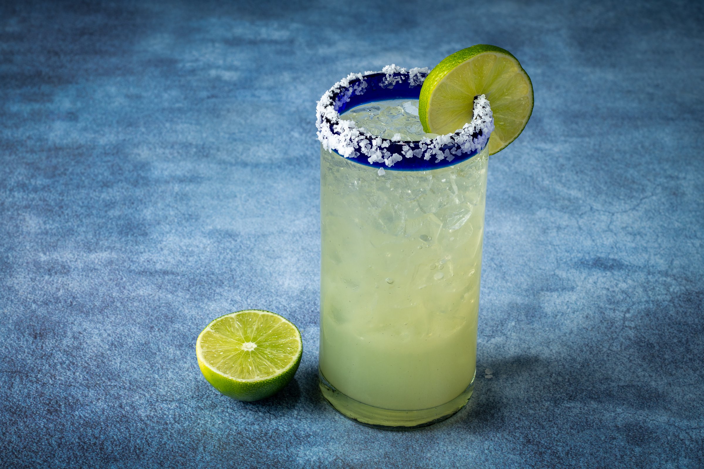 Margarita in a tall glass with a salted rim, a slice of lime on the glass, cut limes in the foreground and a dark background.