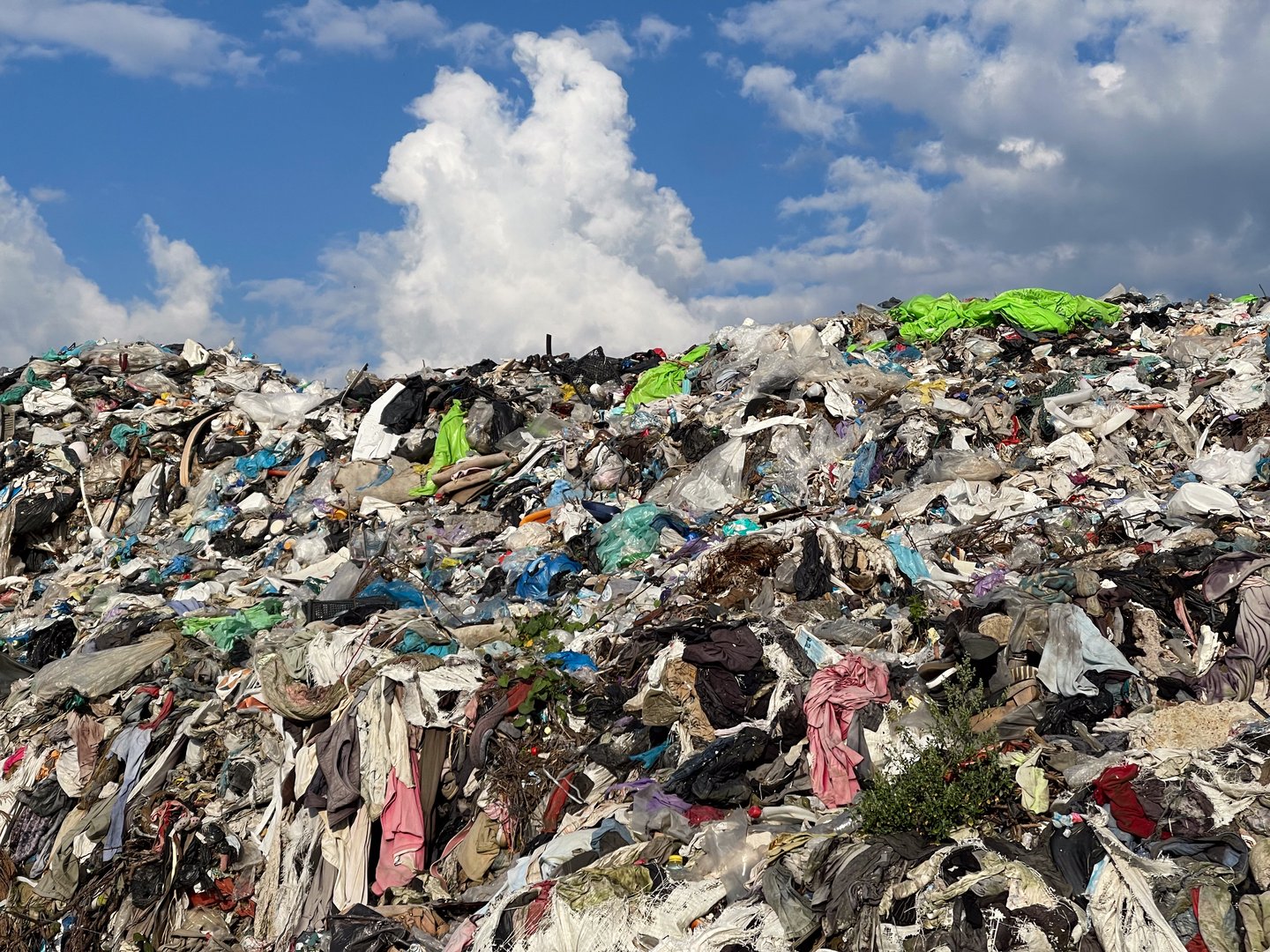 A large landfill displays a mountain of discarded textiles and mixed waste, with vibrant clothing colors contrasting against a blue sky and white clouds