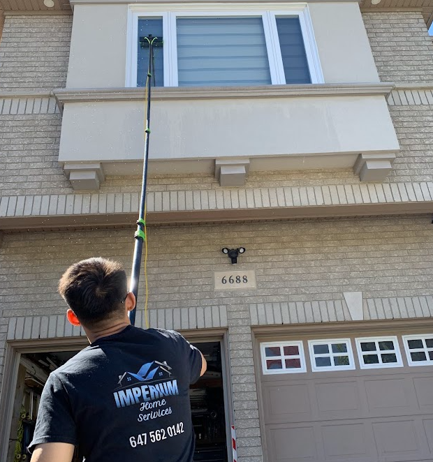 Person cleaning second-floor window of a house using a long pole brush. Wearing a black shirt with a company logo and phone number.