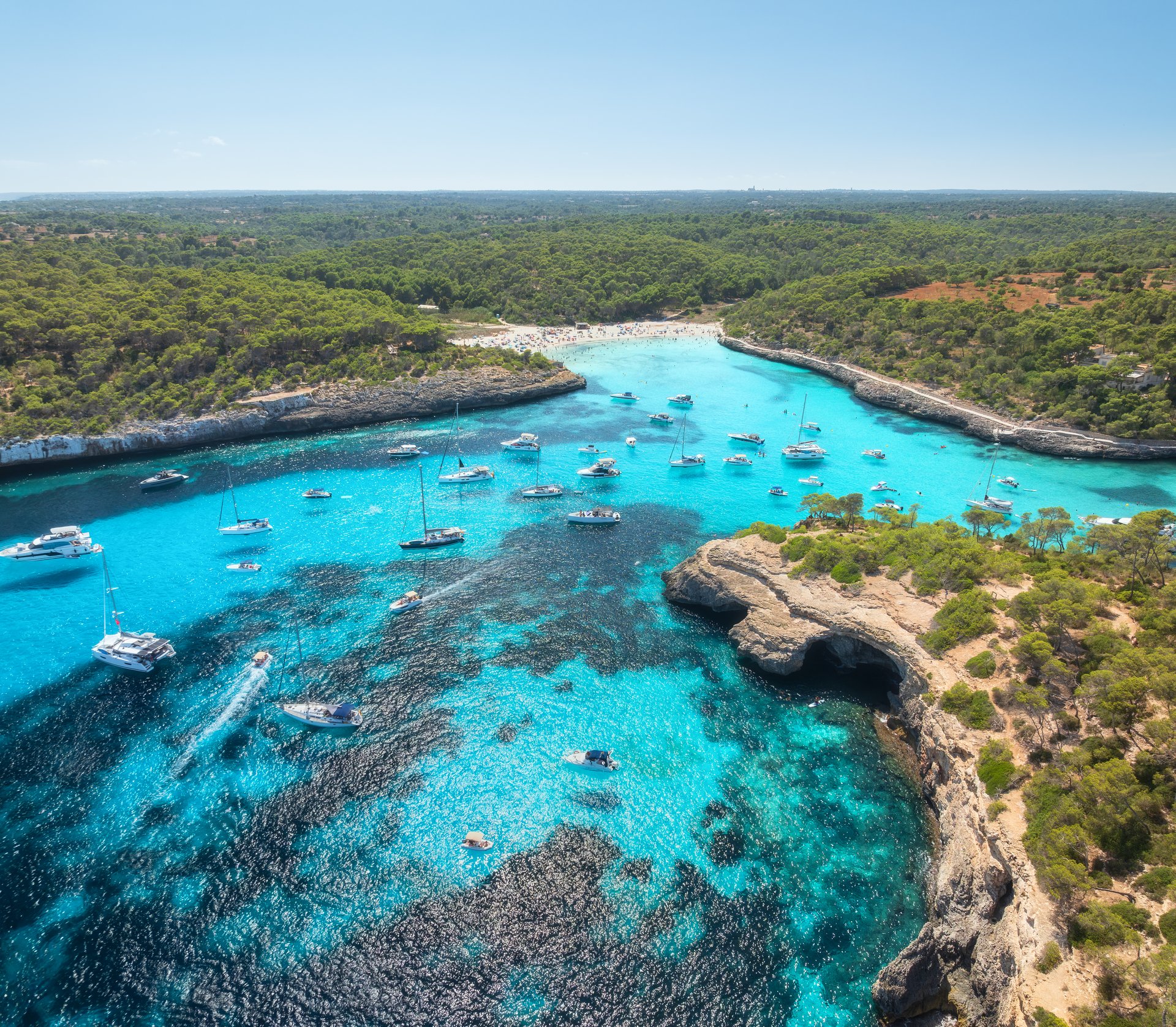 Aerial drone view of yachts and boats anchored in turquoise bay surrounded by rocky cliffs, caves, lush green coastline near Mallorca, Spain. Clear sea, sunny summer day, idyllic seascape. Top view