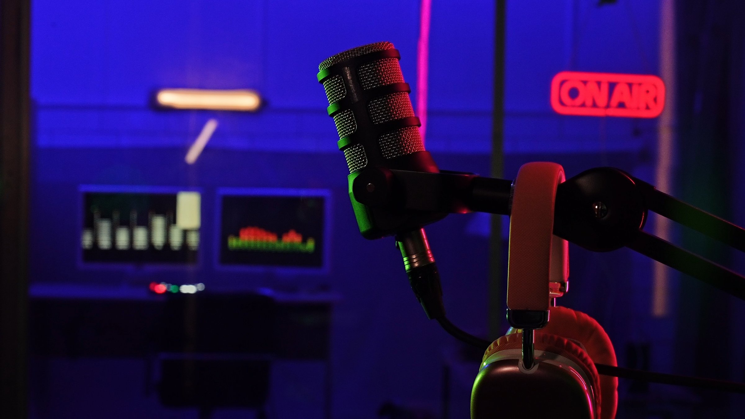 A professional podcast microphone in a neon-lit recording studio. The blurred background features a sound engineer and glowing audio meters, creating a modern broadcasting atmosphere.