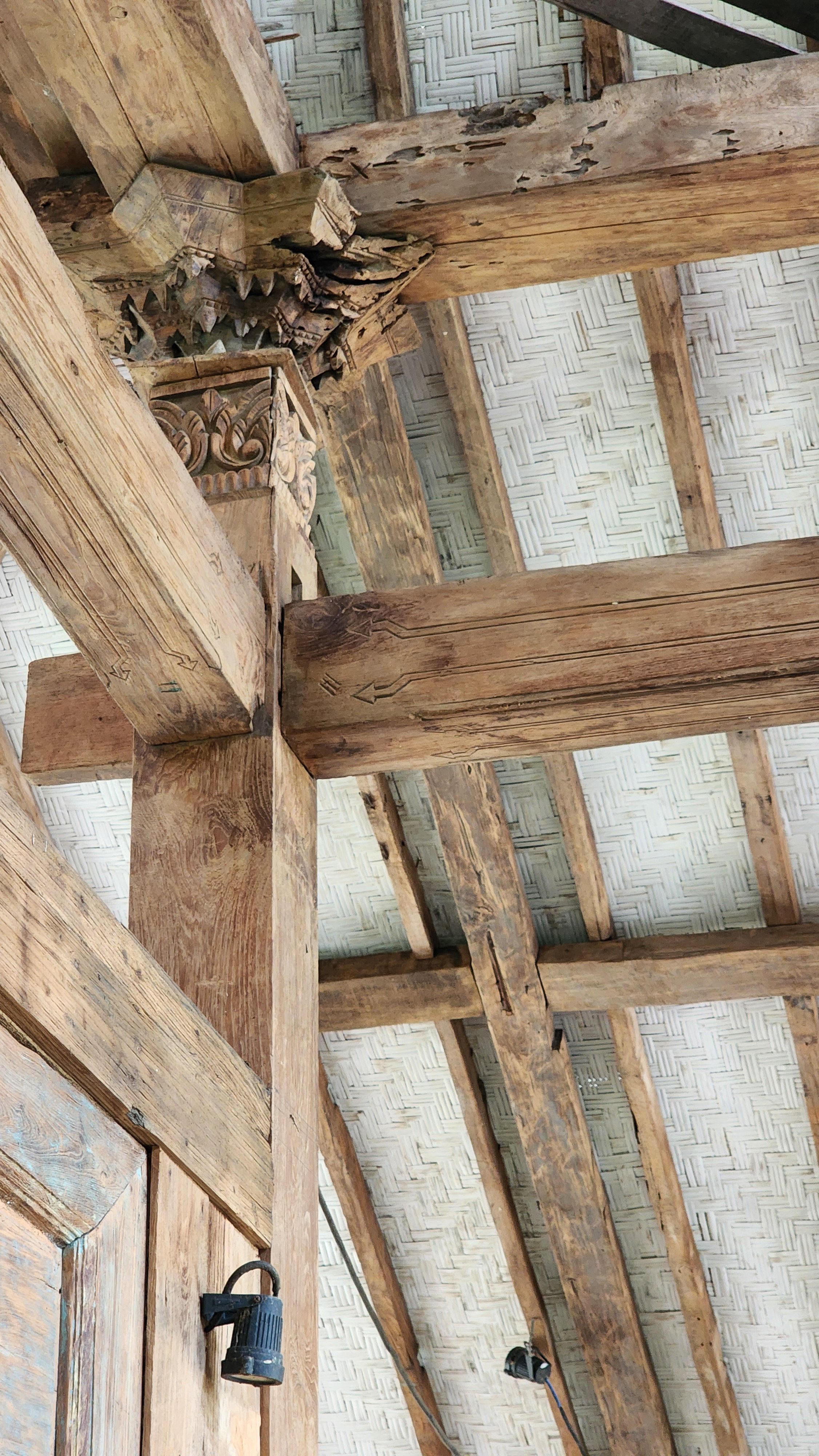 Rustic wooden beams and column support a traditional woven bamboo ceiling, showcasing intricate carvings at the joint.