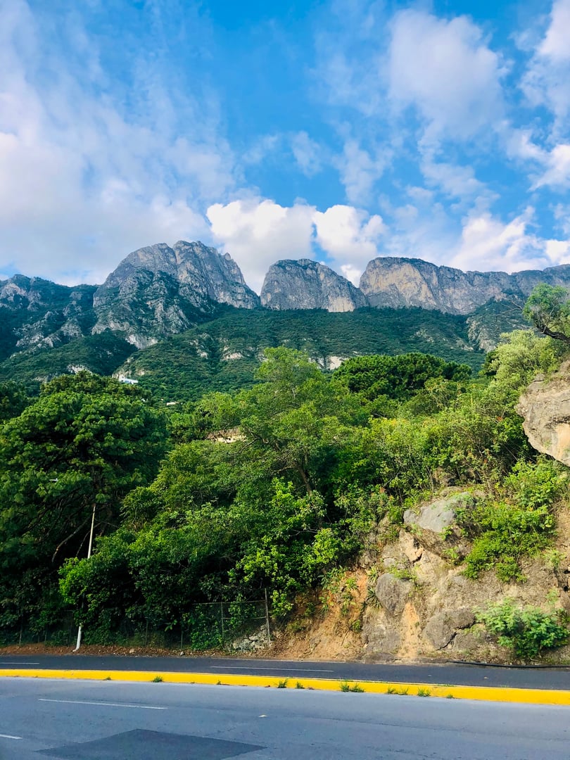 Scenic view of a mountain range under a blue sky with clouds, lush green trees and a road in the foreground.