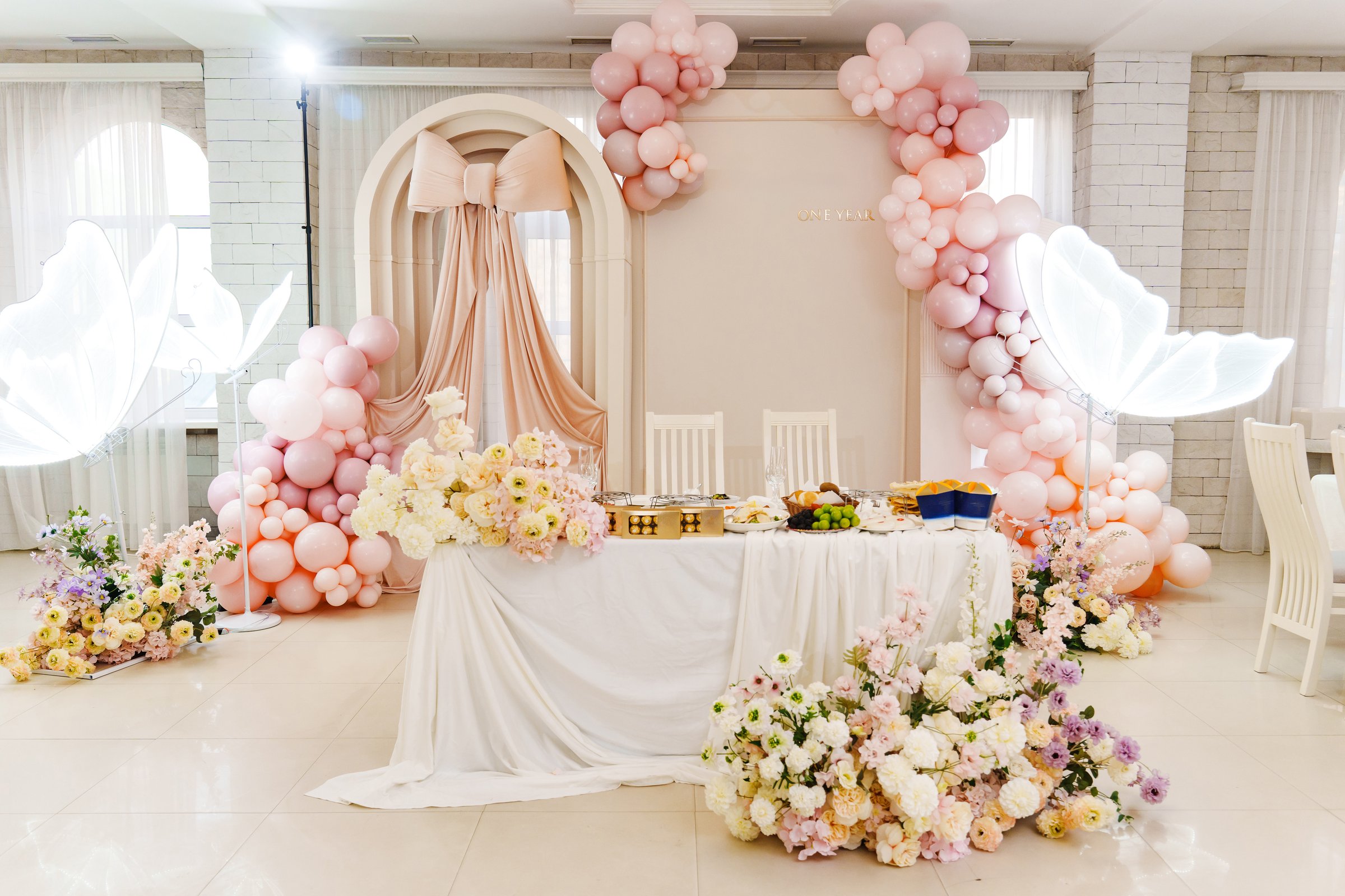 A beautifully decorated event space featuring a table adorned with flowers and pink balloons. Soft lighting enhances the elegant atmosphere, perfect for a special celebration.