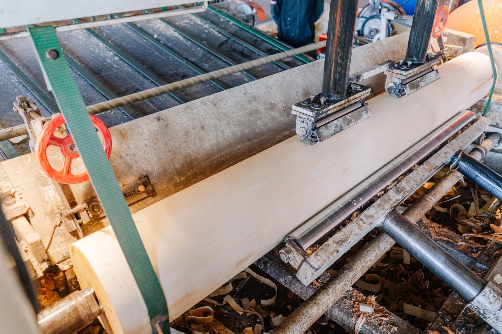A close-up view of a veneer peeling machine processing a large wood log into thin sheets in a woodworking factory. Blade, rollers, and shavings are visible.