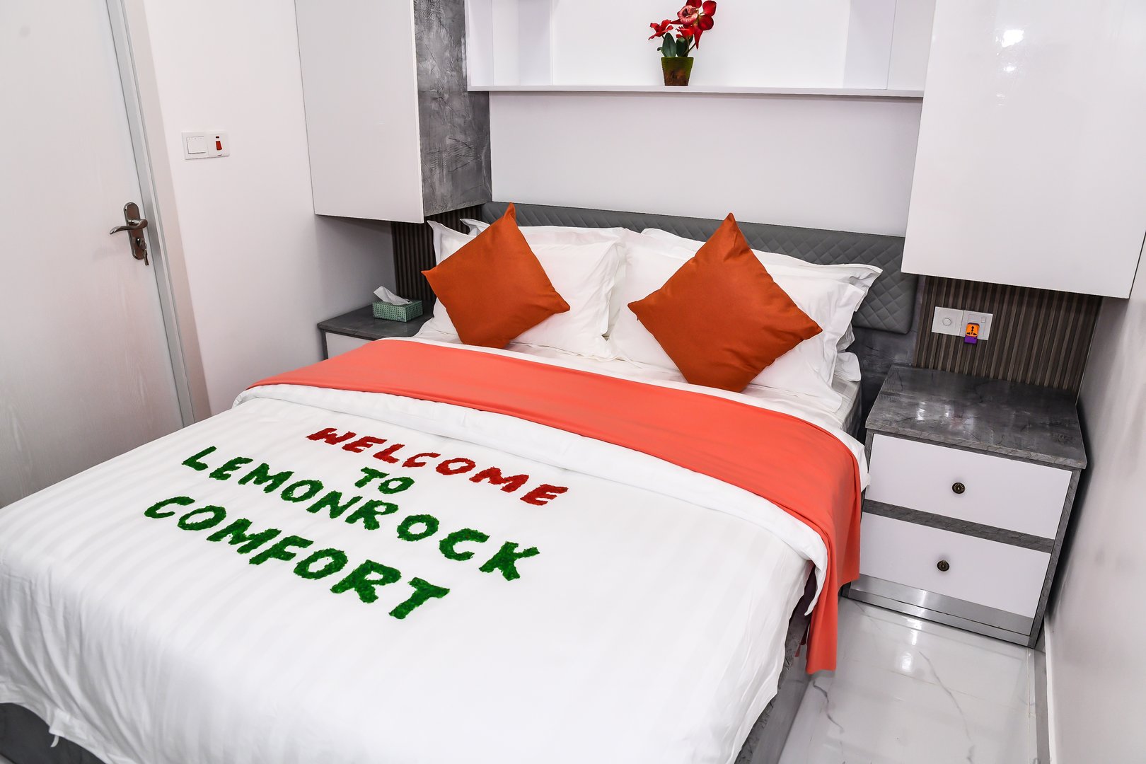 Hotel bed with orange pillows and text Welcome to Lemonrock Comfort written on the white duvet in green and red.