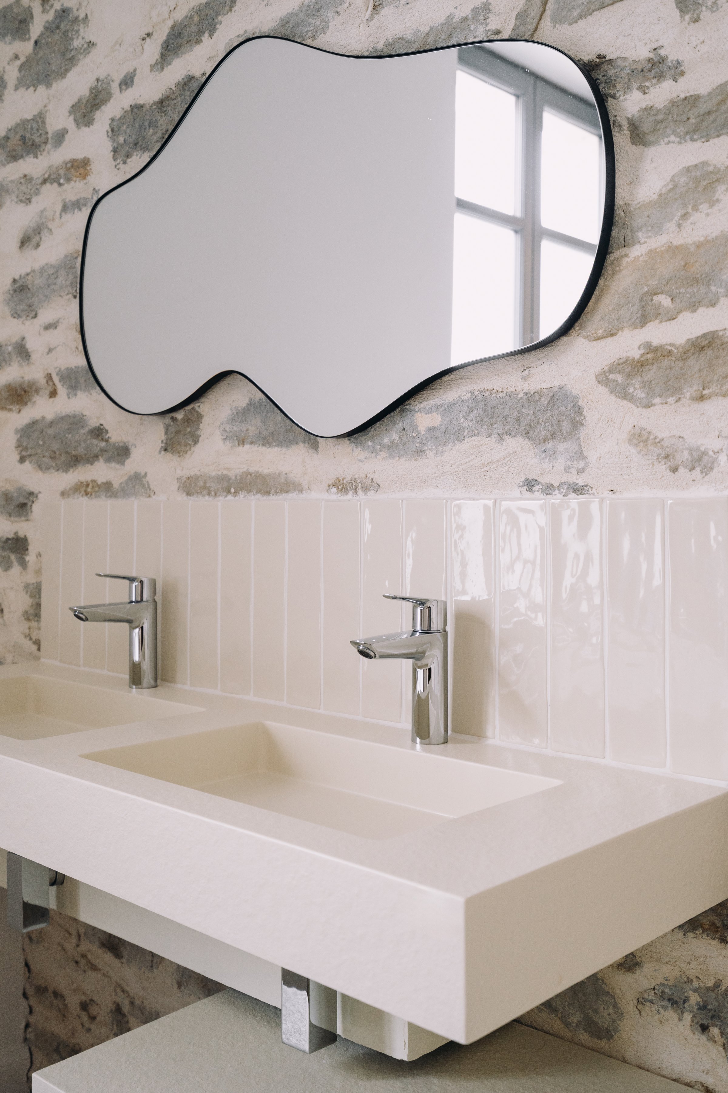 Featuring a contemporary bathroom setup with dual sinks, sleek faucets, and a uniquely shaped mirror against a stone wall, creating a fresh and inviting atmosphere in a residential space.