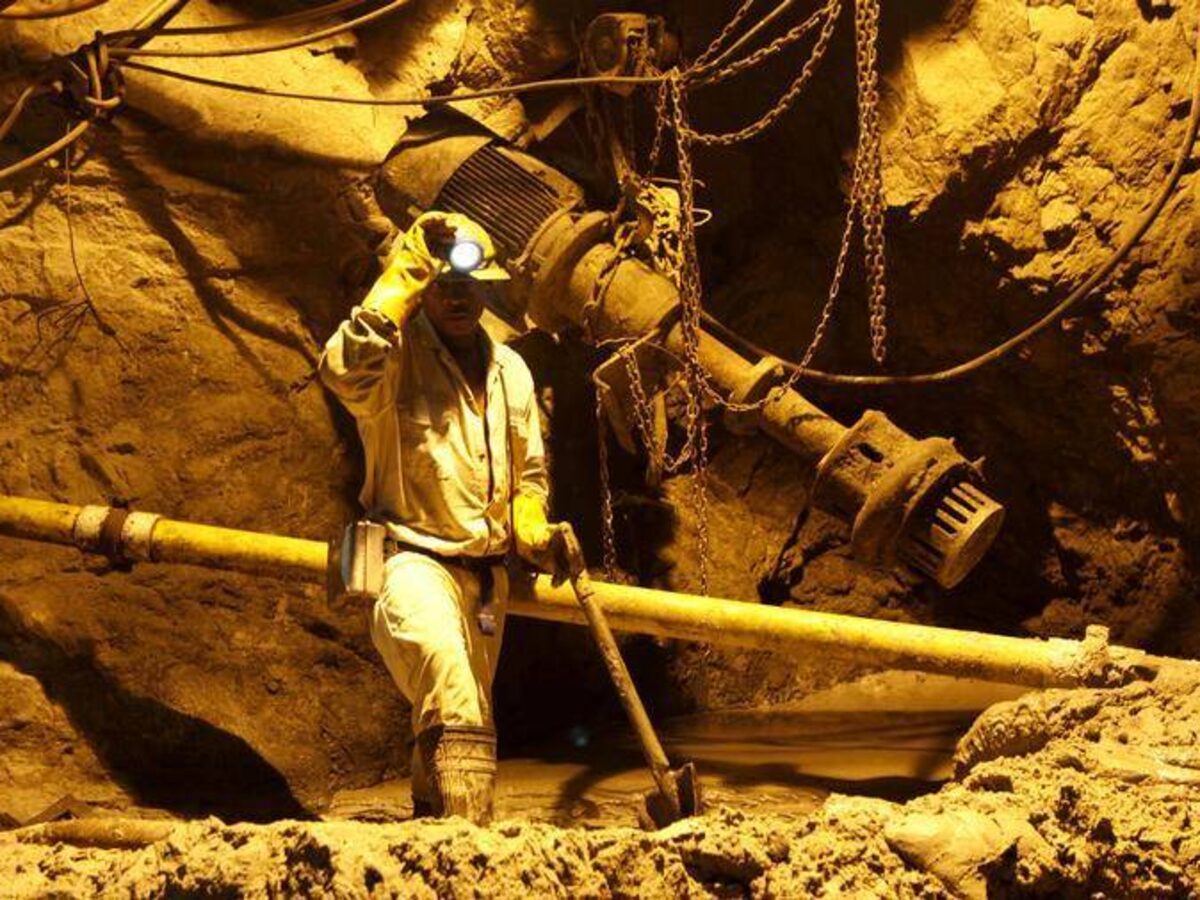 A miner in a dimly lit underground tunnel wearing a helmet with a headlamp and holding a tool, surrounded by equipment and cables.