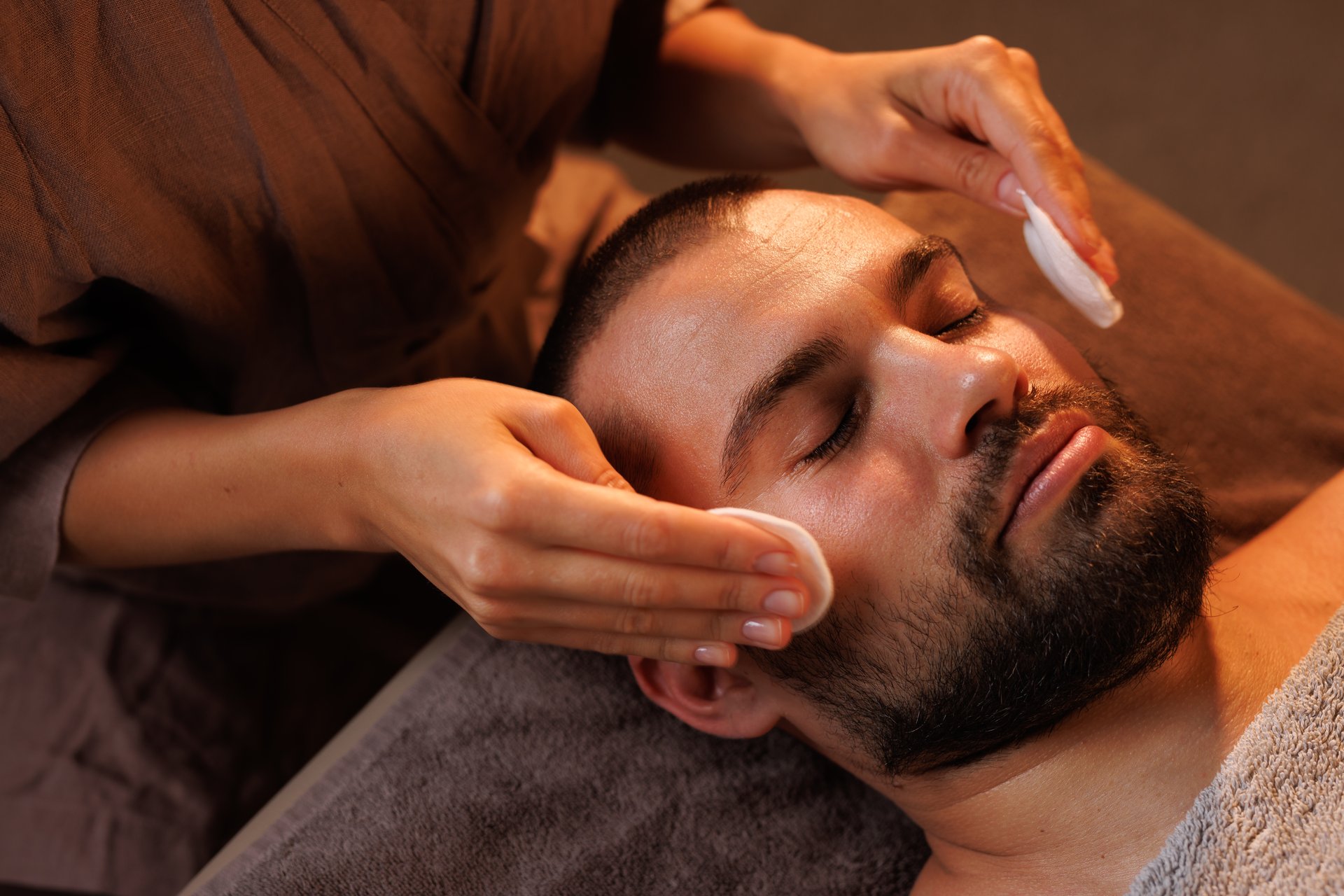 Masseuse wipes man's face with skin sponge, cleansing before facial massage procedure